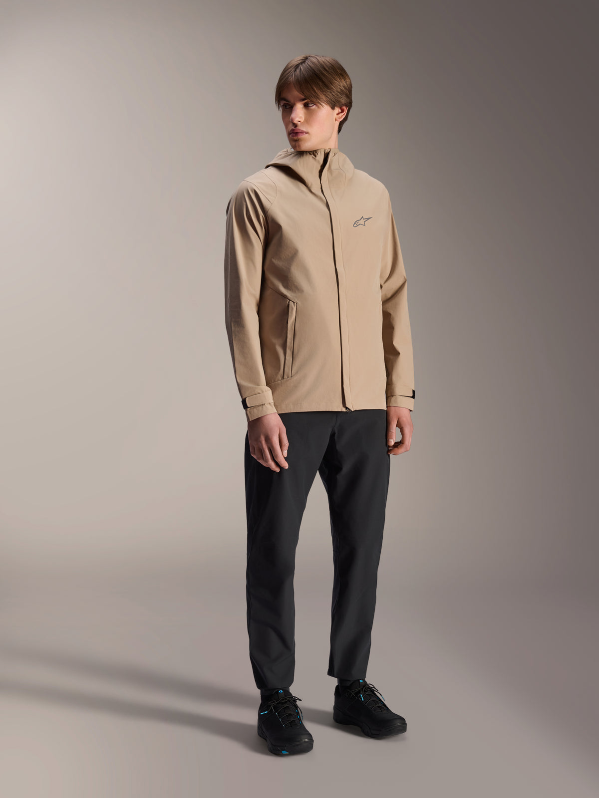 Male model wearing the Alpinestars A-Dura Rain Jacket in Light Brown, paired with black pants and shoes, minimalist outdoor mountain bike rain jacket featuring a hooded design and a small chest logo.