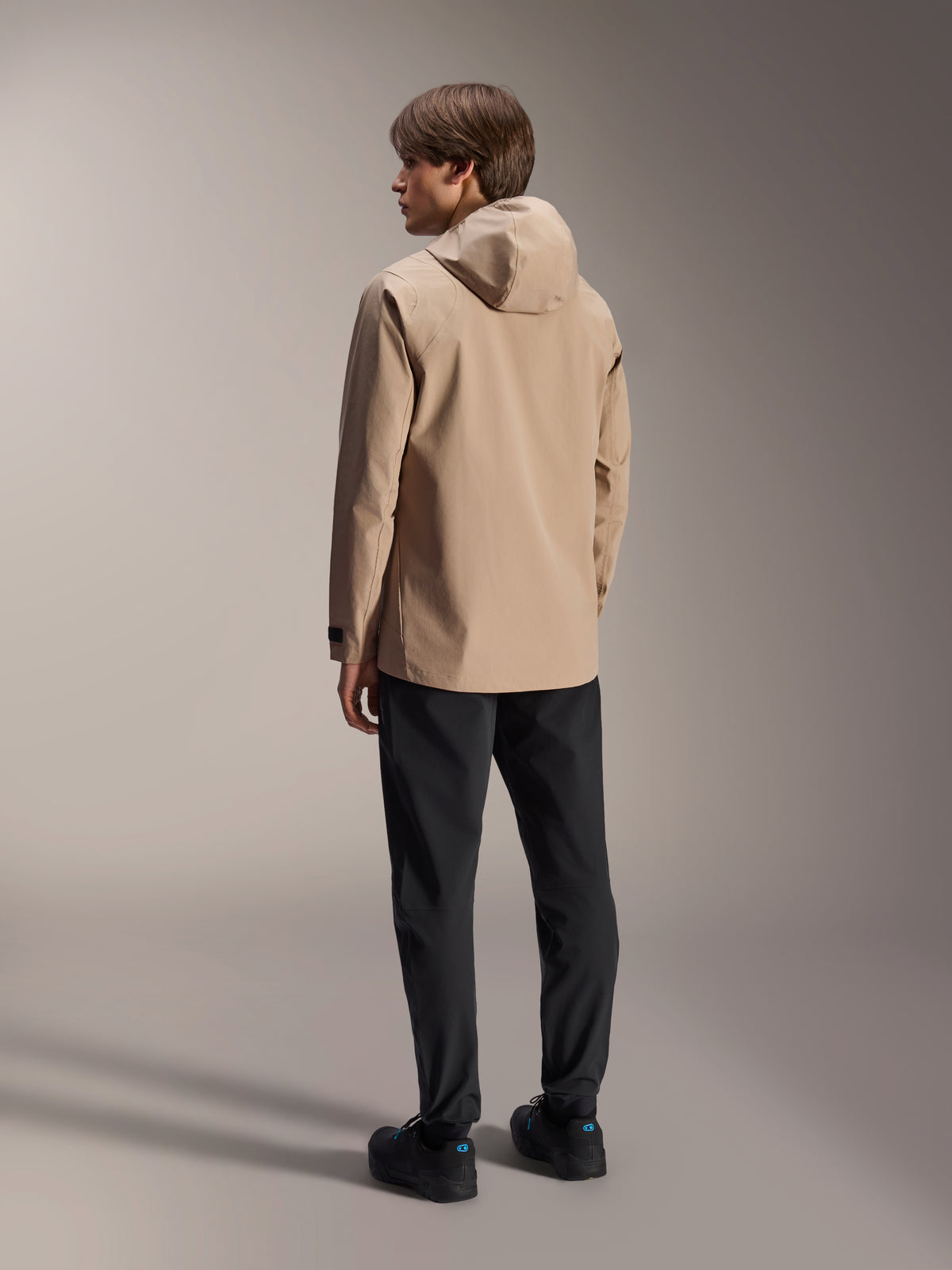 Alpinestars A-Dura Rain Jacket, Light Brown, performance MTB outerwear, model viewed from behind wearing the jacket with hood, paired with black pants and black shoes, technical design for all-weather mountain biking