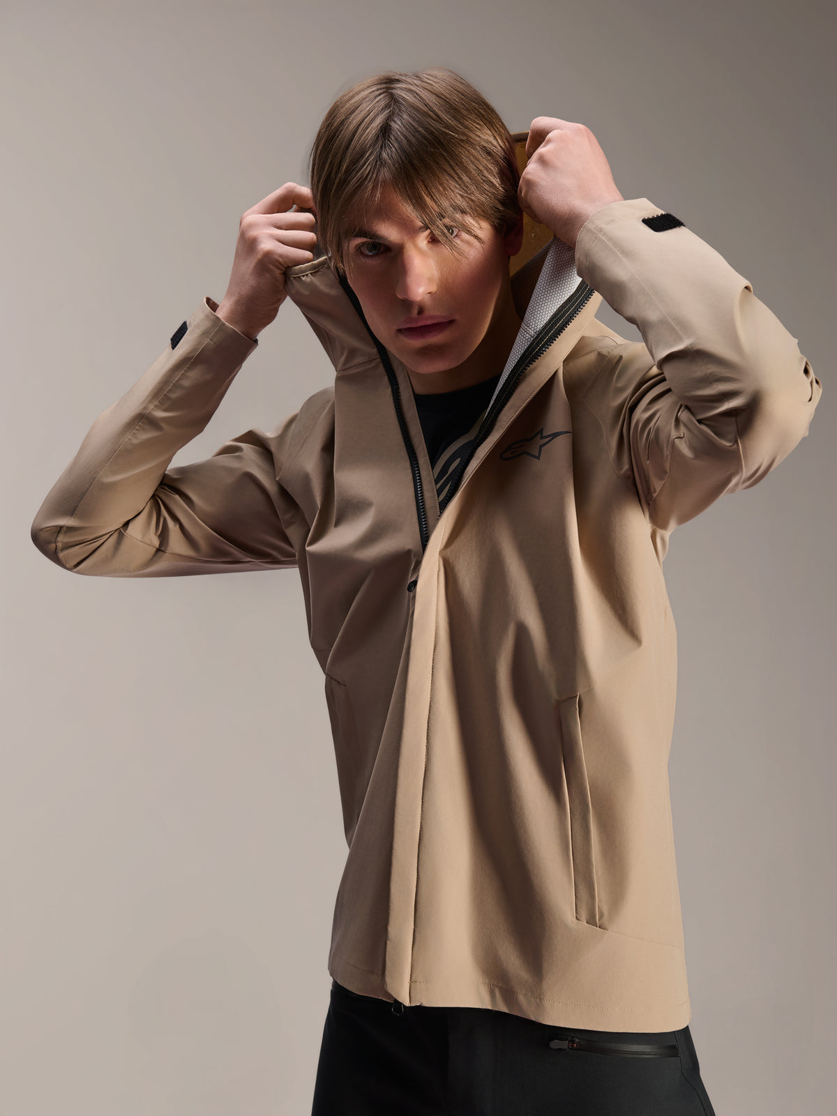 Alpinestars A-Dura Rain Jacket, MTB Cycling Jacket, Light Brown, model wearing the jacket and pulling up the elasticated hood, featuring a black chest logo, zippered pockets, and technical 2.5-layer waterproof fabric
