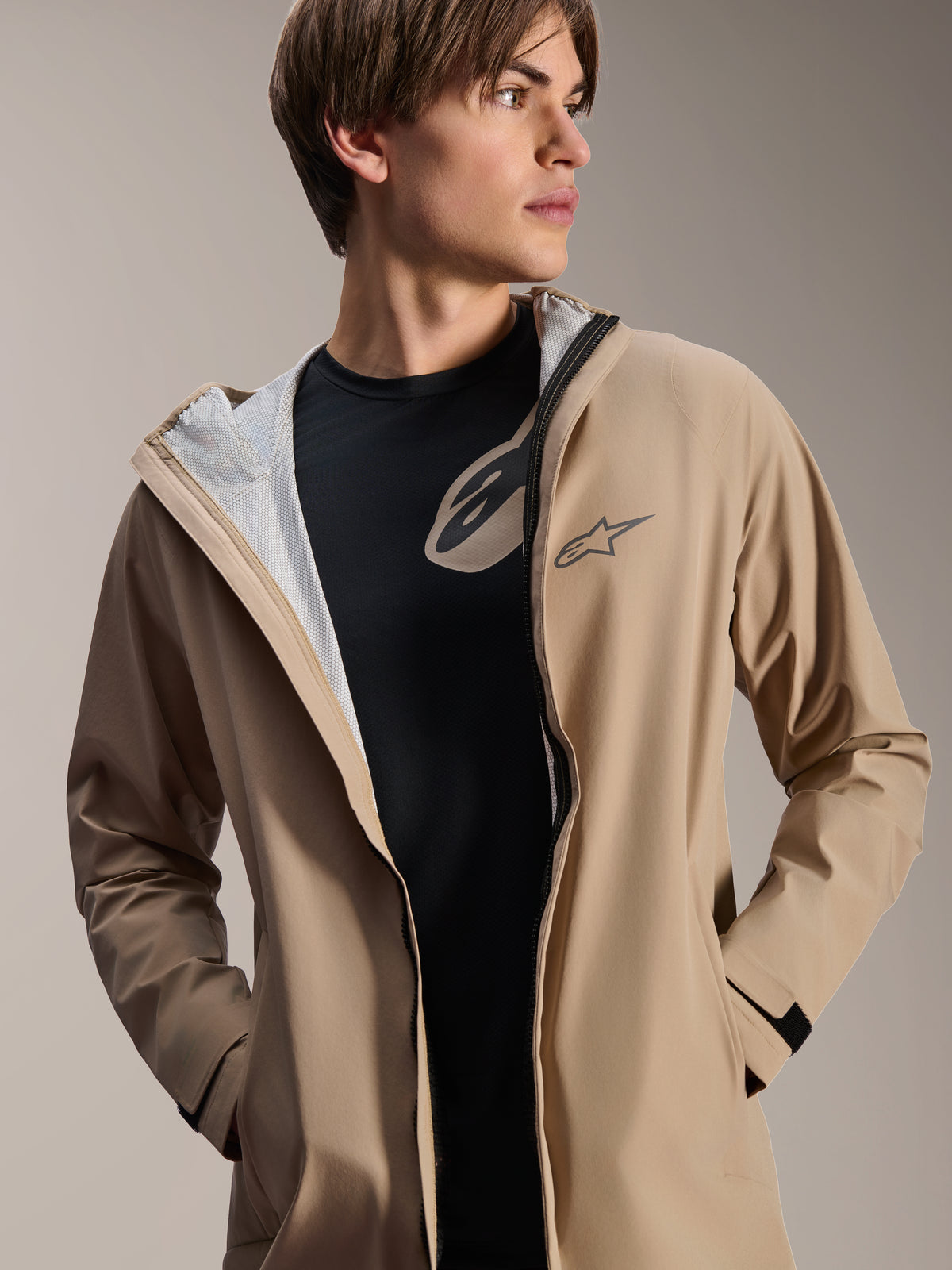 Male model wearing Alpinestars A-Dura Rain Jacket, Light Brown, unzipped over a black t-shirt, showing white mesh lining, versatile weather-resistant cycling jacket