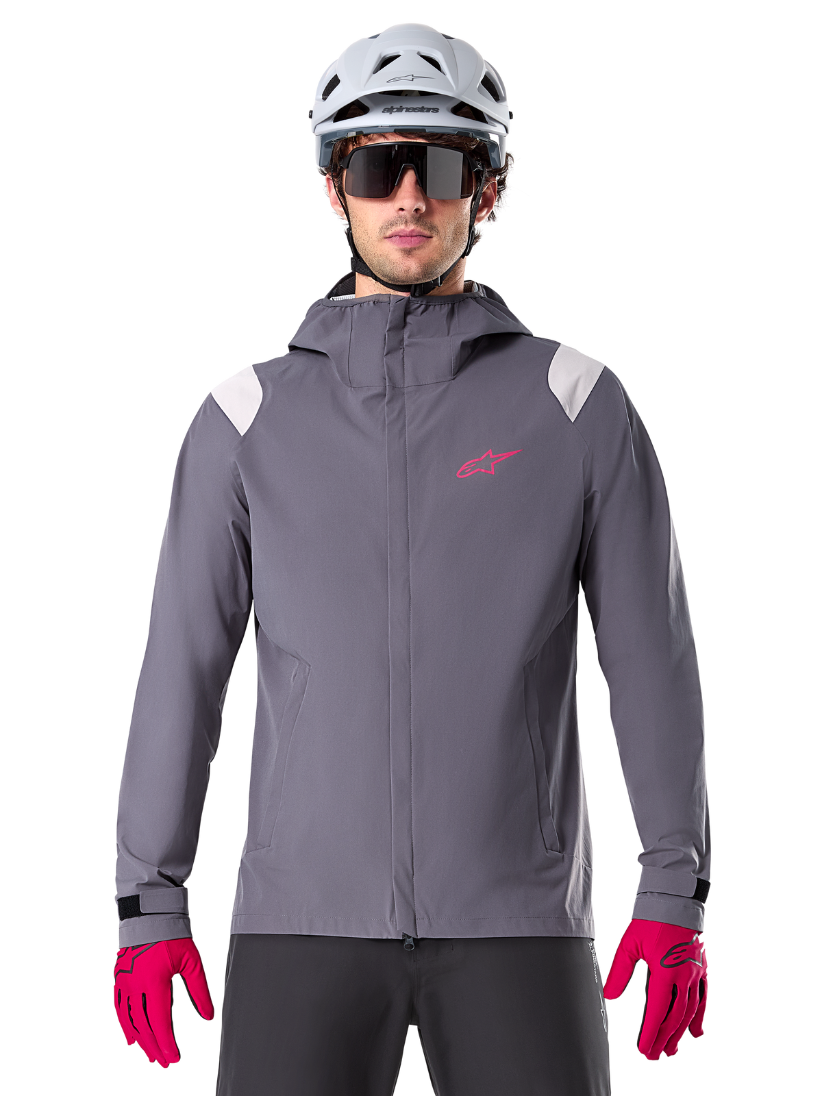 Veste de pluie Alpinestars A-Dura, veste de pluie pour VTT, gris titanium foncé, modèle à capuche avec logo Alpinestars rose et empiècements gris clair aux épaules, portée avec un casque de VTT blanc et des gants rose vif, équipement imperméable pour le cyclisme tout-terrain