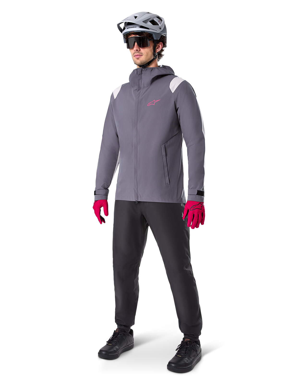 Modèle masculin portant la veste de pluie Alpinestars A-Dura en Titanium Foncé, veste grise à capuche avec logo rose sur la poitrine et empiècements gris clair aux épaules, complétée par un casque de VTT gris, des lunettes de soleil, des gants rose vif et un pantalon noir pour le cyclisme tout-terrain