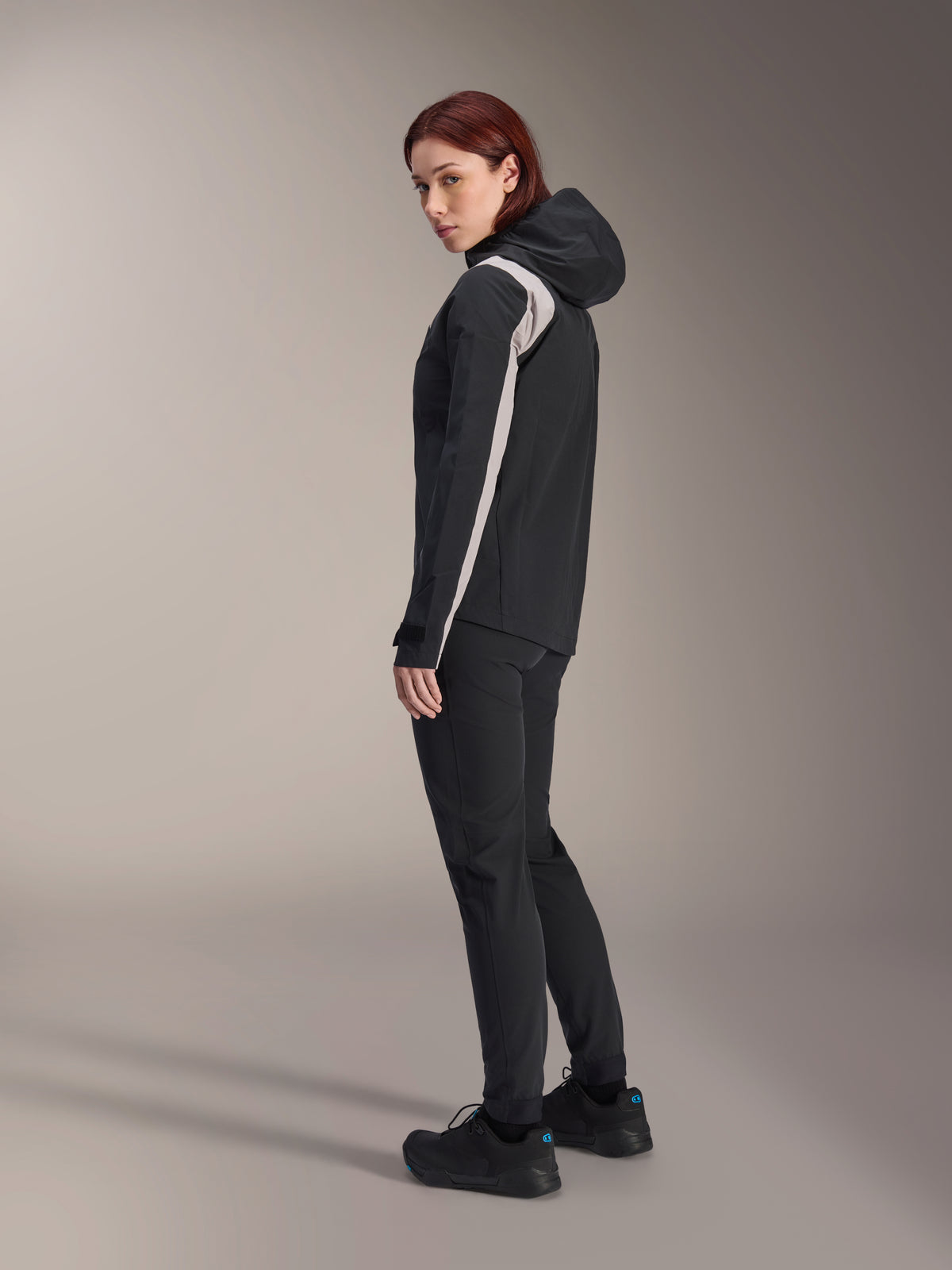 Woman wearing Alpinestars Stella A-Dura Rain Jacket, Black with light gray stripe on the sleeve, hooded waterproof cycling jacket for MTB, paired with black technical pants and black riding shoes, studio profile view