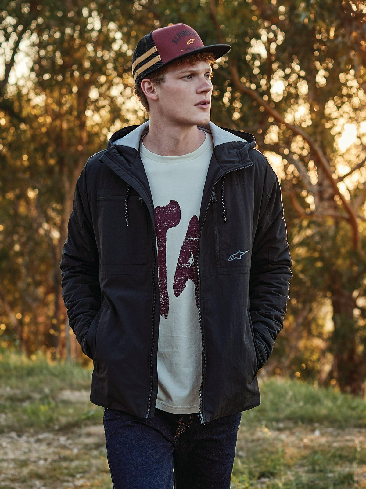Casquette Trucker Backline Alpinestars en brique, noir et moutarde, portée par un homme avec une veste à capuche noire et un t-shirt graphique, look streetwear décontracté inspiré du sport automobile en plein air à l'heure dorée.