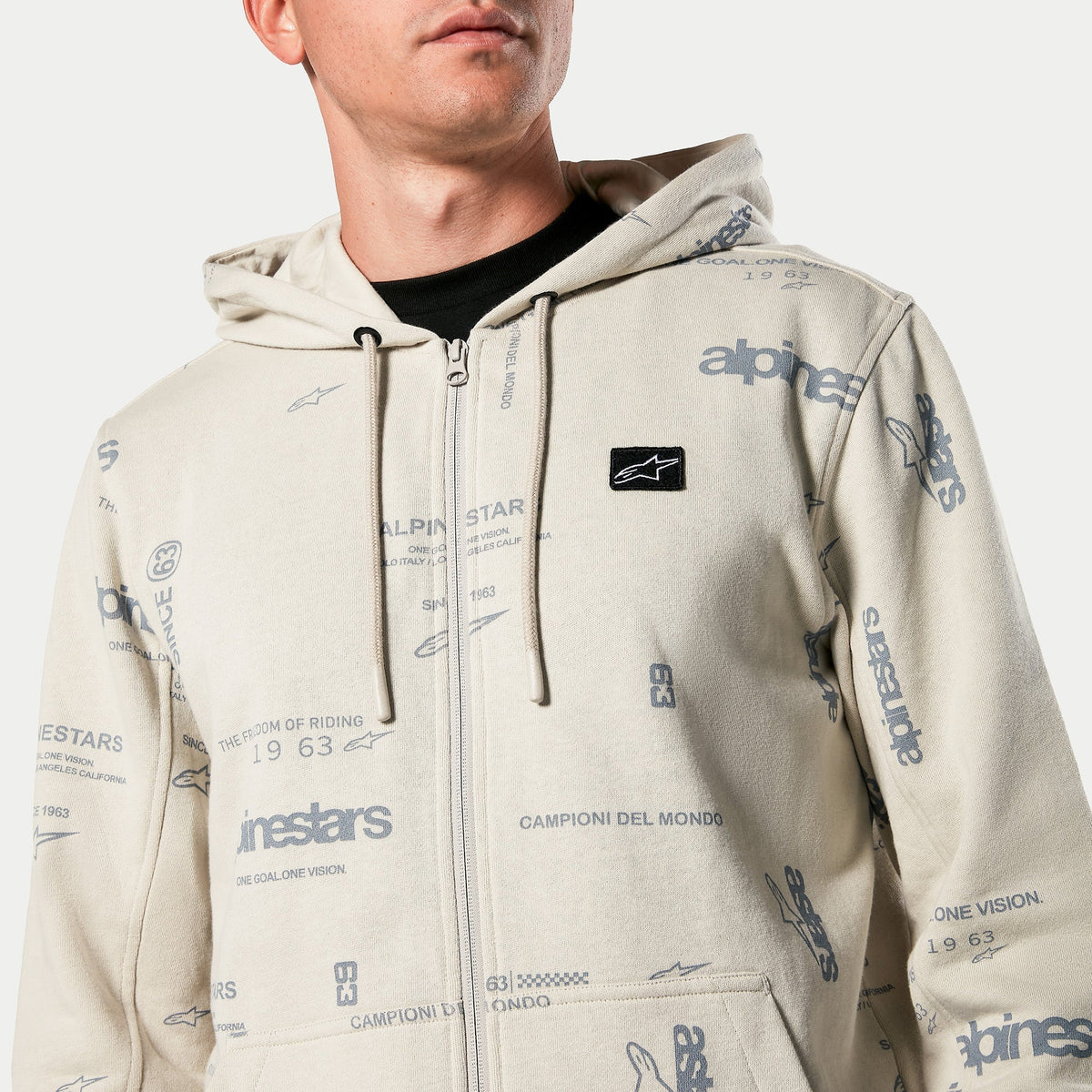 Sweat à capuche Alpinestars Plenitude, polaire zippée en blanc cassé, imprimé graphique intégral avec textes « alpinestars » et « 1963 », patch logo noir brodé sur la poitrine, capuche à cordon de serrage, style streetwear décontracté inspiré du sport automobile