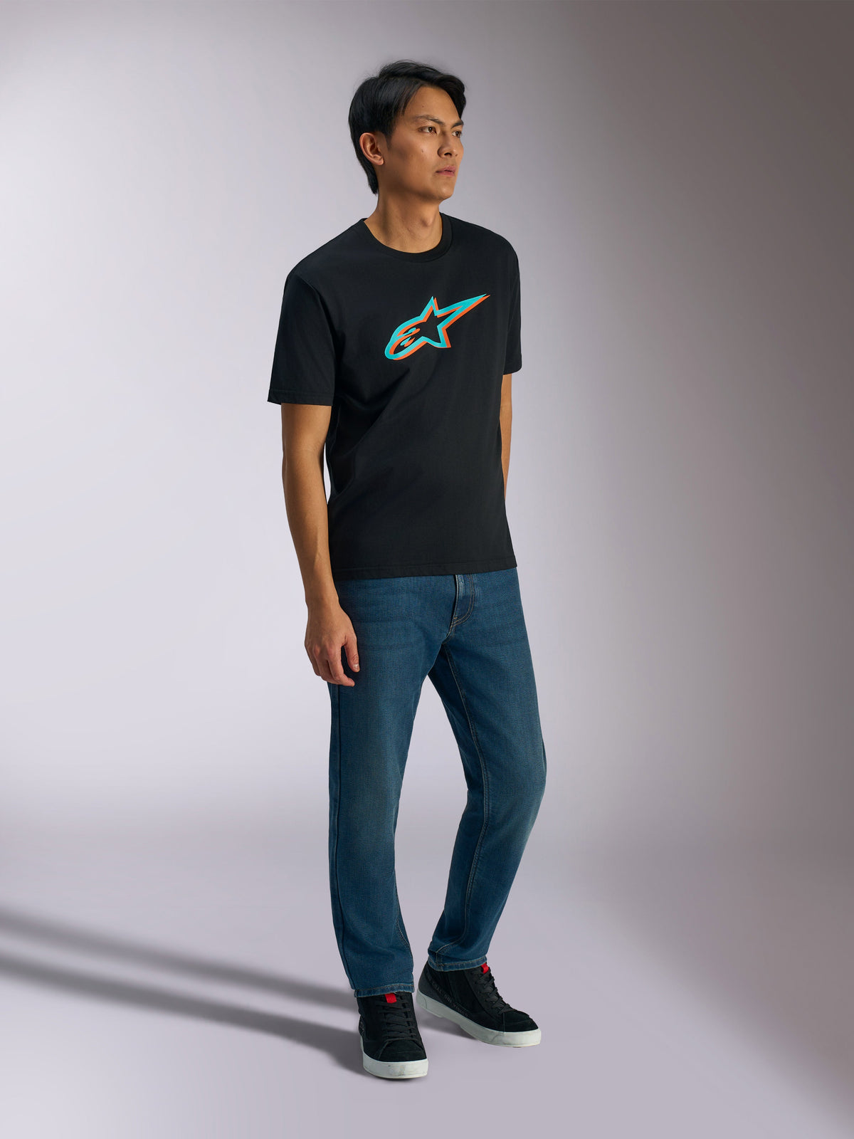Alpinestars Ageless Shadow CSF Tee, t-shirt à manches courtes, noir avec logo graphique effet ombre turquoise et orange, porté par un modèle masculin avec un jean bleu et des baskets noires, streetwear décontracté d'inspiration motorsport