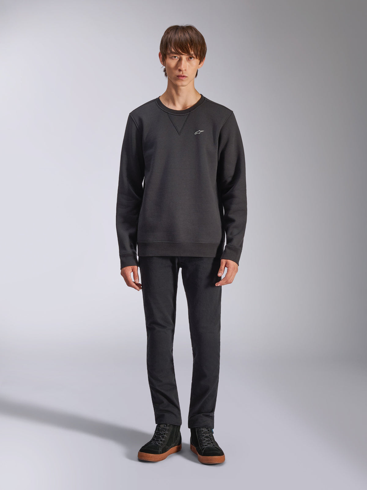 Sweatshirt Alpinestars Positive Crew, pull à col rond noir décontracté, petit logo blanc sur la poitrine, encolure avec surpiqûre en V, photo de studio en pied avec un mannequin portant un pantalon noir et des baskets, vêtements lifestyle d'inspiration motorsport