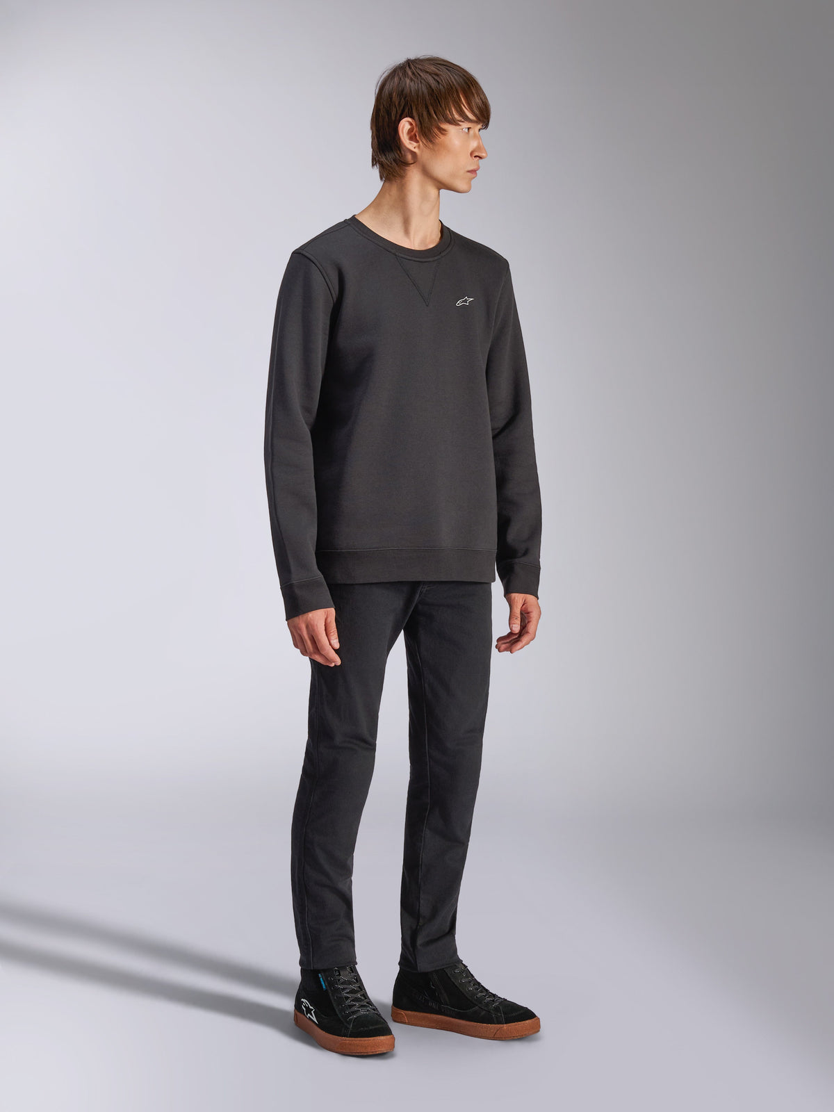 Alpinestars Positive Crew, sweat-shirt à col rond, noir, petit logo Alpinestars blanc sur la poitrine, col côtelé avec détail de couture en V, poignets et ourlet côtelés, porté par un mannequin avec un pantalon noir et des baskets, streetwear décontracté inspiré du sport automobile