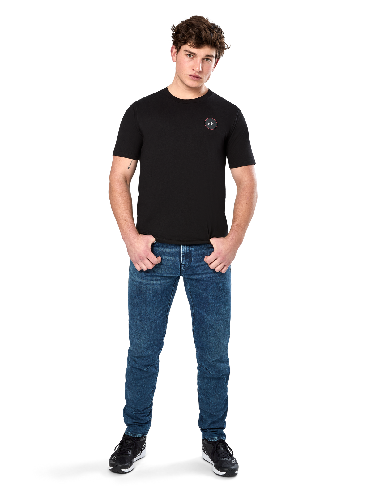 Alpinestars Dot Carbon CSF Tee, t-shirt à manches courtes, noir, petit logo circulaire Astars Dot sur la poitrine, porté avec un jean bleu et des baskets noires, style streetwear décontracté inspiré du sport automobile