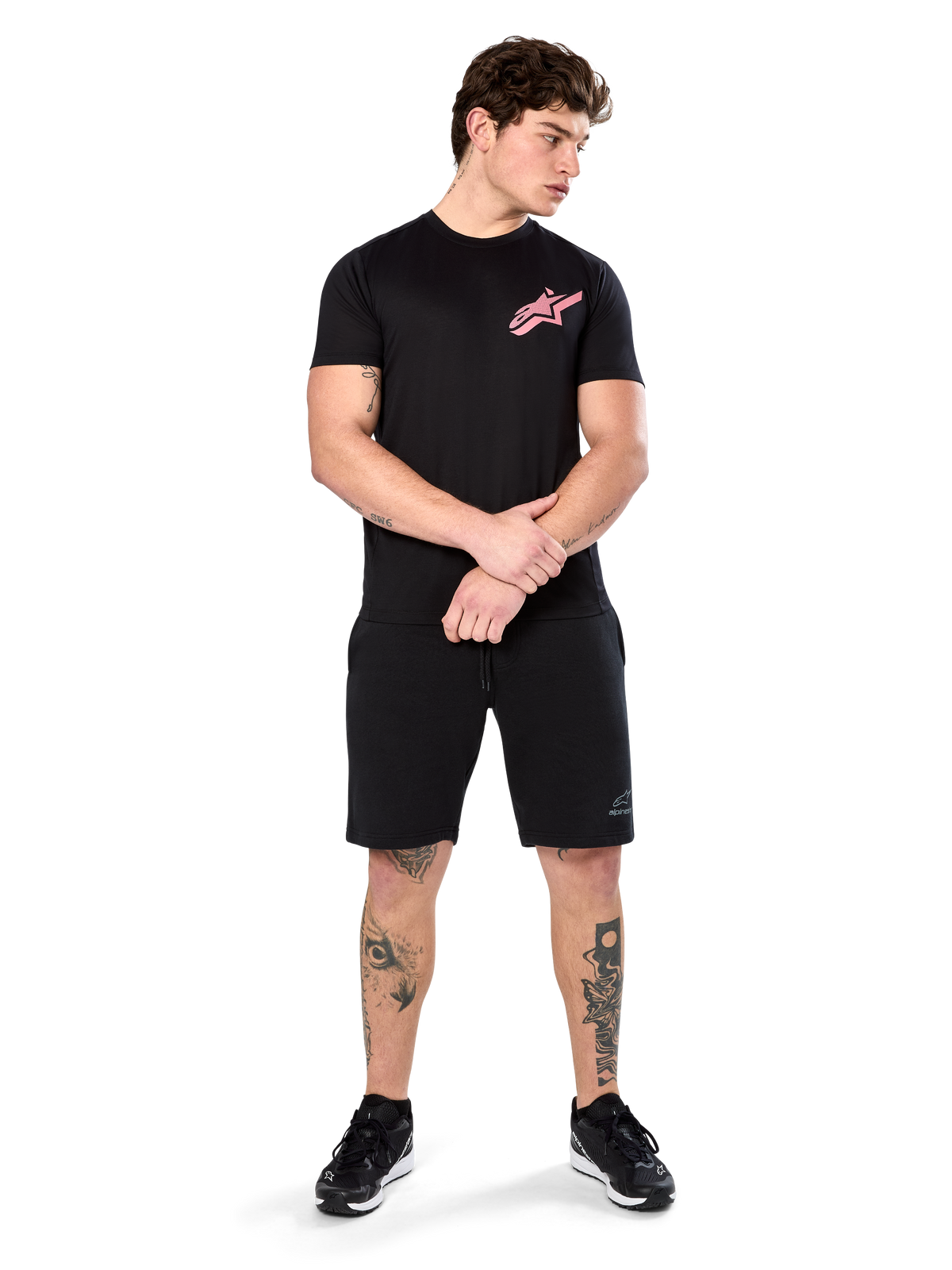 Modèle portant le t-shirt Alpinestars Attrition Performance, t-shirt de performance à manches courtes, noir avec un logo Astars rose sur la poitrine, assorti à un short noir et des baskets, vêtement de sport décontracté inspiré du sport automobile