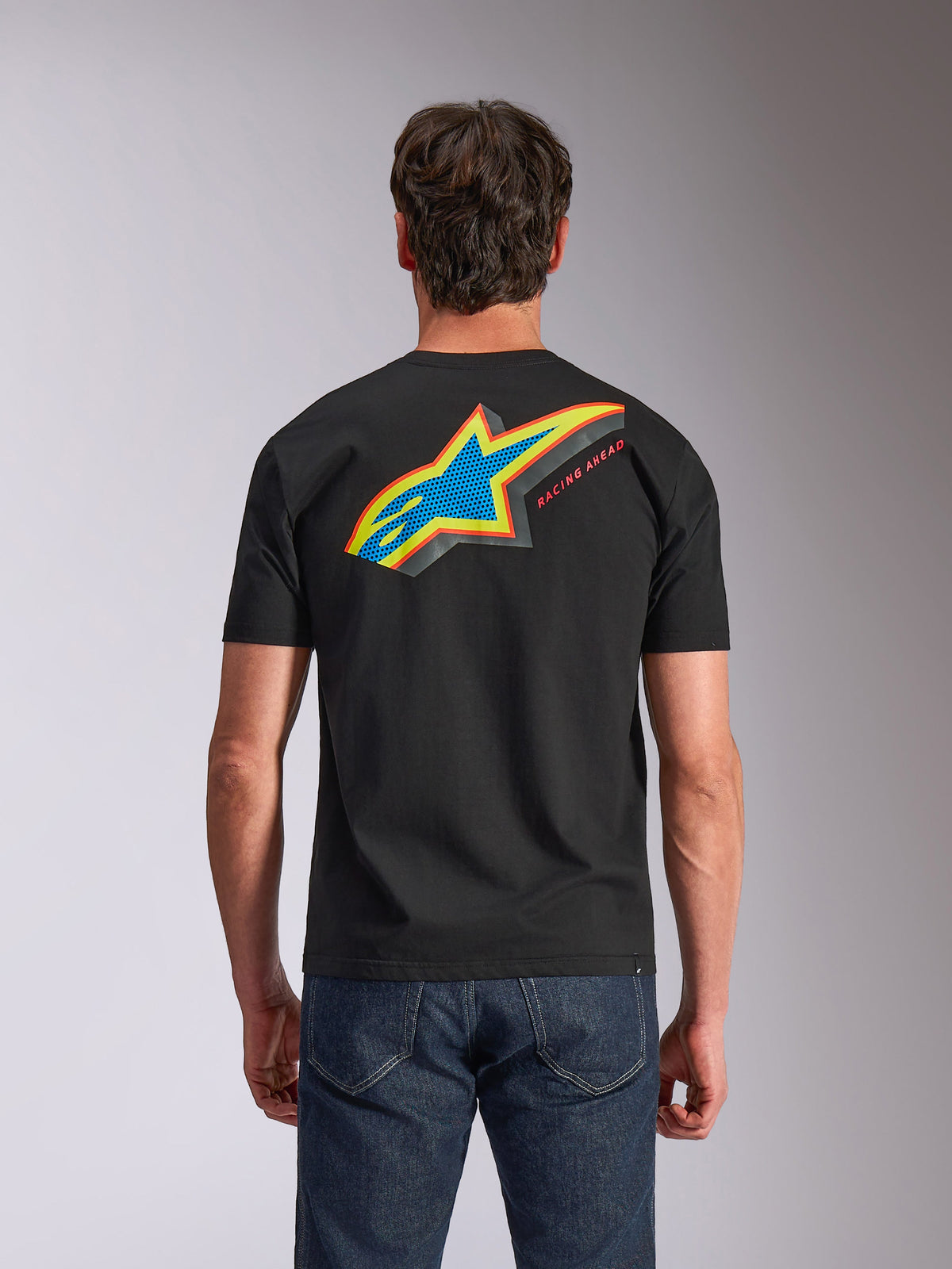 Alpinestars Angler CSF Tee, t-shirt à manches courtes, noir, vue de dos montrant le logo Astars multicolore vibrant avec remplissage perforé et le texte « RACING AHEAD », porté avec un jean bleu, vêtement décontracté d'inspiration sport automobile