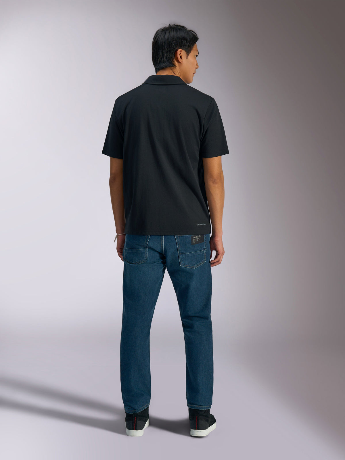 Polo Sport Impetus Alpinestars, noir, vue arrière d'un polo à manches courtes avec marquage Alpinestars dans le bas du dos, porté avec un jean bleu, vêtement sportswear décontracté inspiré du sport automobile