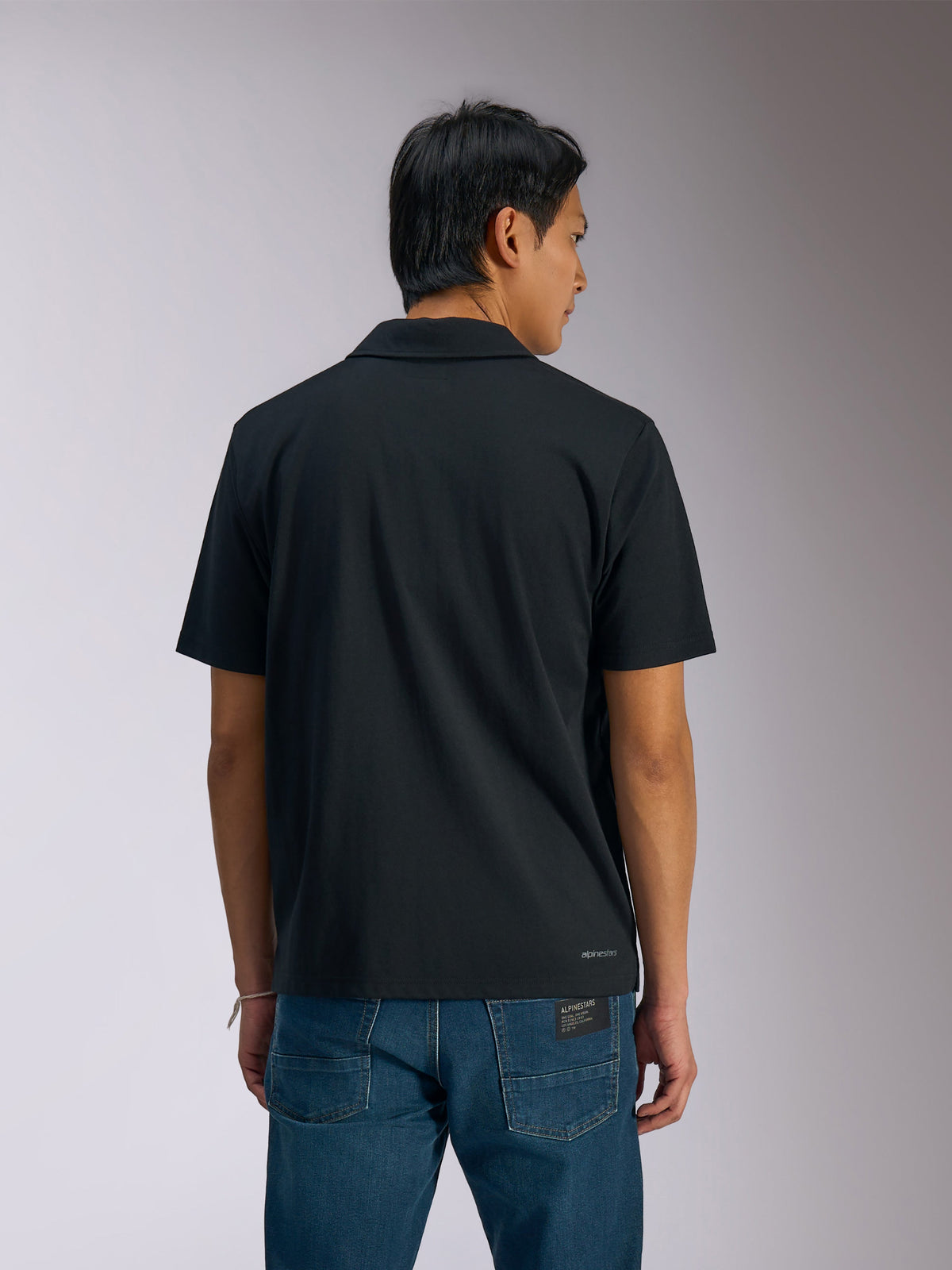 Polo Sport Impetus Alpinestars, polo noir à manches courtes, vue de dos d'un homme portant le polo avec un jean denim bleu, présentant un petit marquage Alpinestars en bas du dos, vêtement sportswear décontracté inspiré du sport automobile