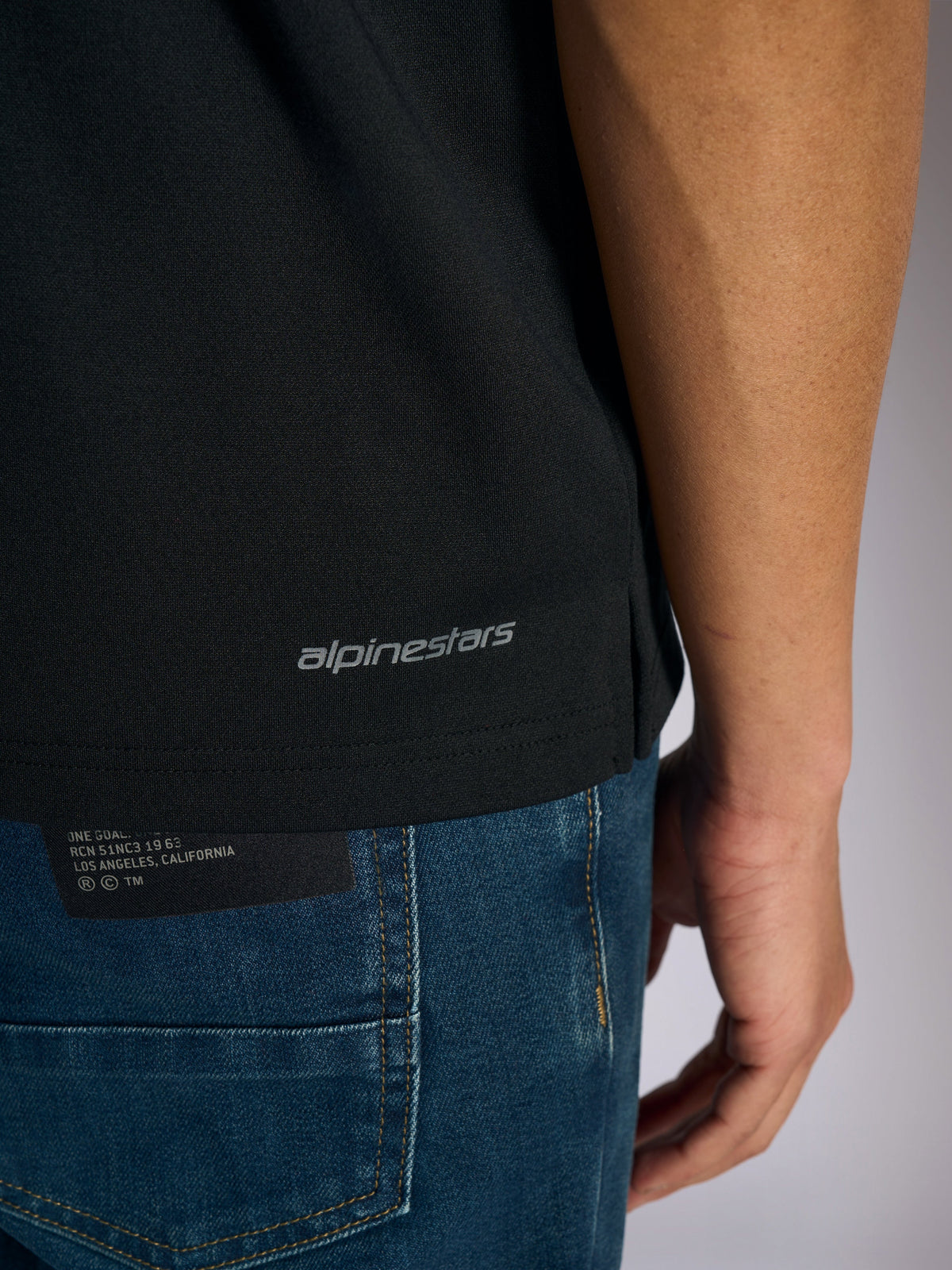Polo Alpinestars Impetus Sport, vue détaillée de l'ourlet inférieur et de la fente latérale, noir avec marquage Alpinestars gris, sportswear décontracté inspiré du sport automobile