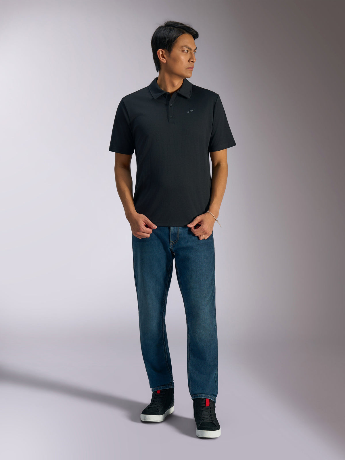 Polo Sport Alpinestars Impetus, polo noir à manches courtes avec patte de boutonnage à trois boutons et logo Astars gris sur la poitrine, porté par un homme avec un jean bleu et des baskets montantes noires, vêtement de sport décontracté d'inspiration sport automobile