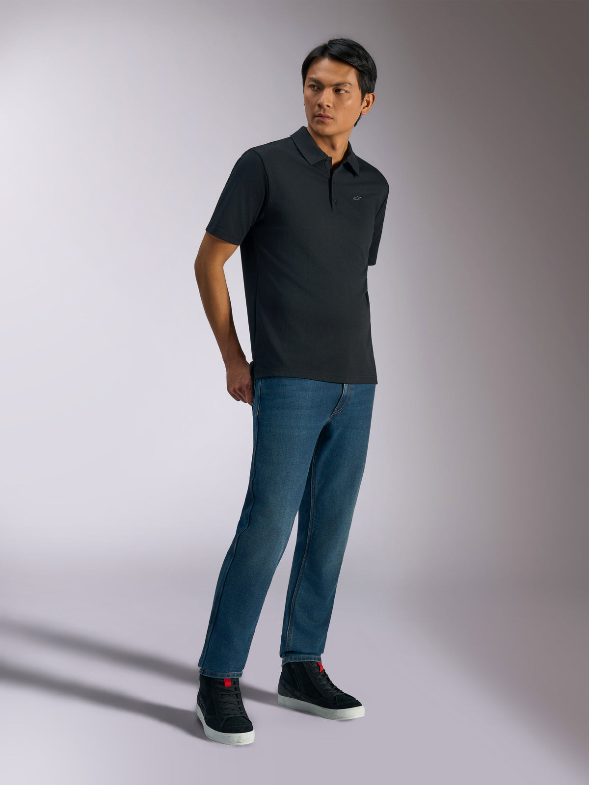 Polo Sport Impetus Alpinestars, polo à manches courtes, noir, doté d'un logo discret sur la poitrine et d'une patte de boutonnage à trois boutons, porté par un mannequin avec un jean bleu et des baskets noires, vêtement lifestyle décontracté inspiré du sport automobile