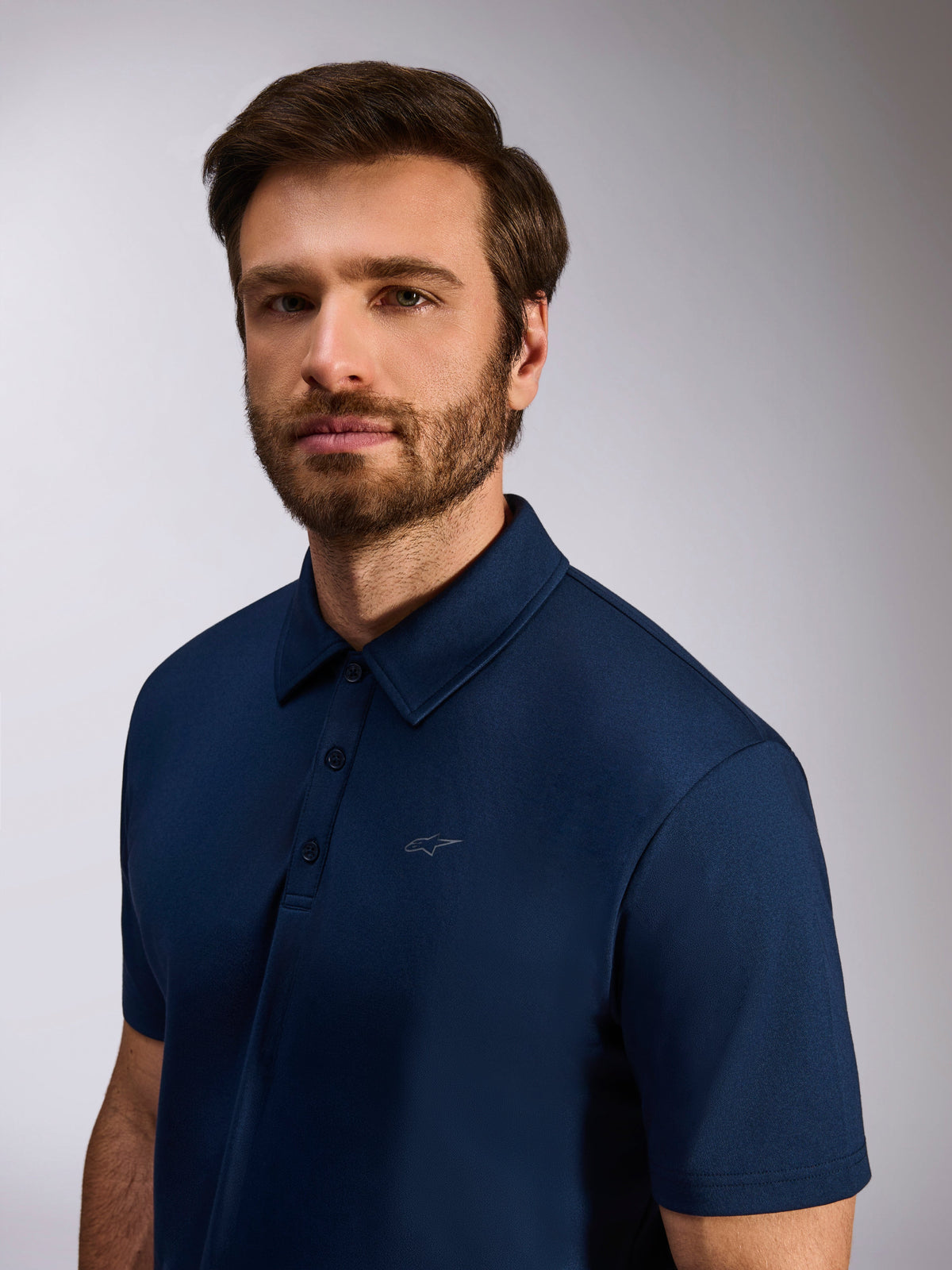 Modèle portant le Polo Sport Impetus d'Alpinestars, bleu marine, polo à manches courtes en maille imitation avec patte à trois boutons et discret logo Astars sur la poitrine, vêtement de style motorsport décontracté