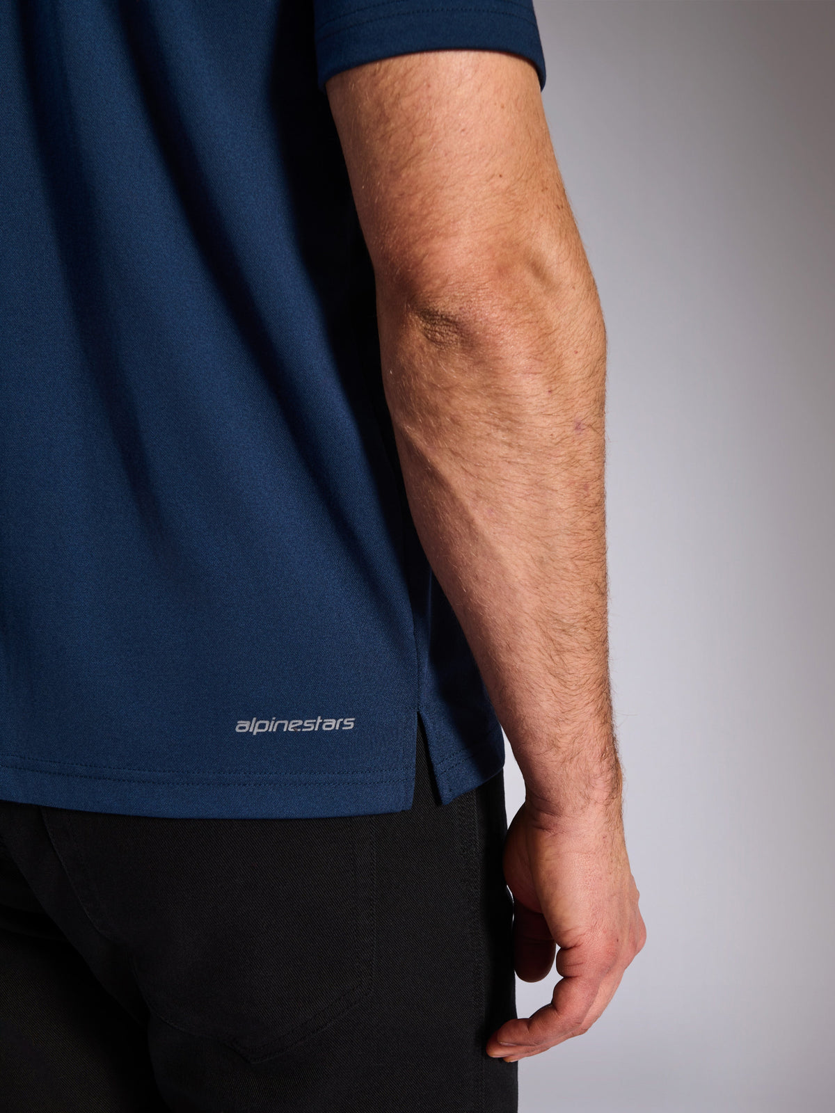 Polo Sport Impetus Alpinestars, bleu marine, gros plan sur l'ourlet inférieur montrant le logo Alpinestars et la fente latérale, texture en maille imitation, vêtement de sport décontracté inspiré du motorsport