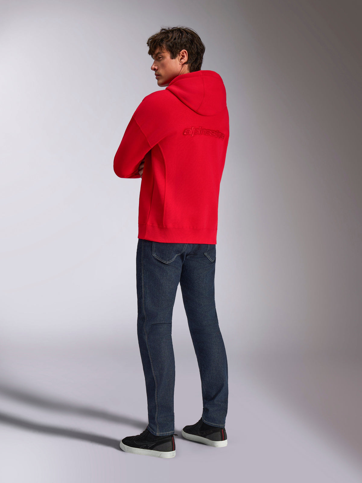 Sweat à capuche oversized Alpinestars Emergent, rouge, vue de dos montrant le logo Alpinestars brodé au dos, coupe ample, porté avec un jean bleu foncé et des baskets noires, style streetwear décontracté