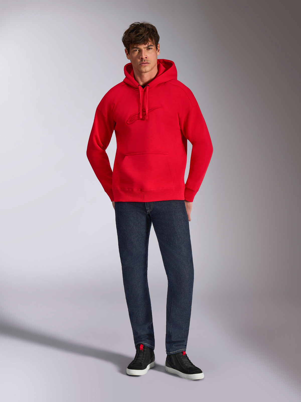 Sweat à capuche Alpinestars Emergent Oversized, rouge, arborant un logo Astars brodé en 3D ton sur ton sur la poitrine, poche kangourou et capuche à cordon de serrage, porté par un mannequin avec un jean bleu foncé et des baskets noires, style streetwear décontracté.