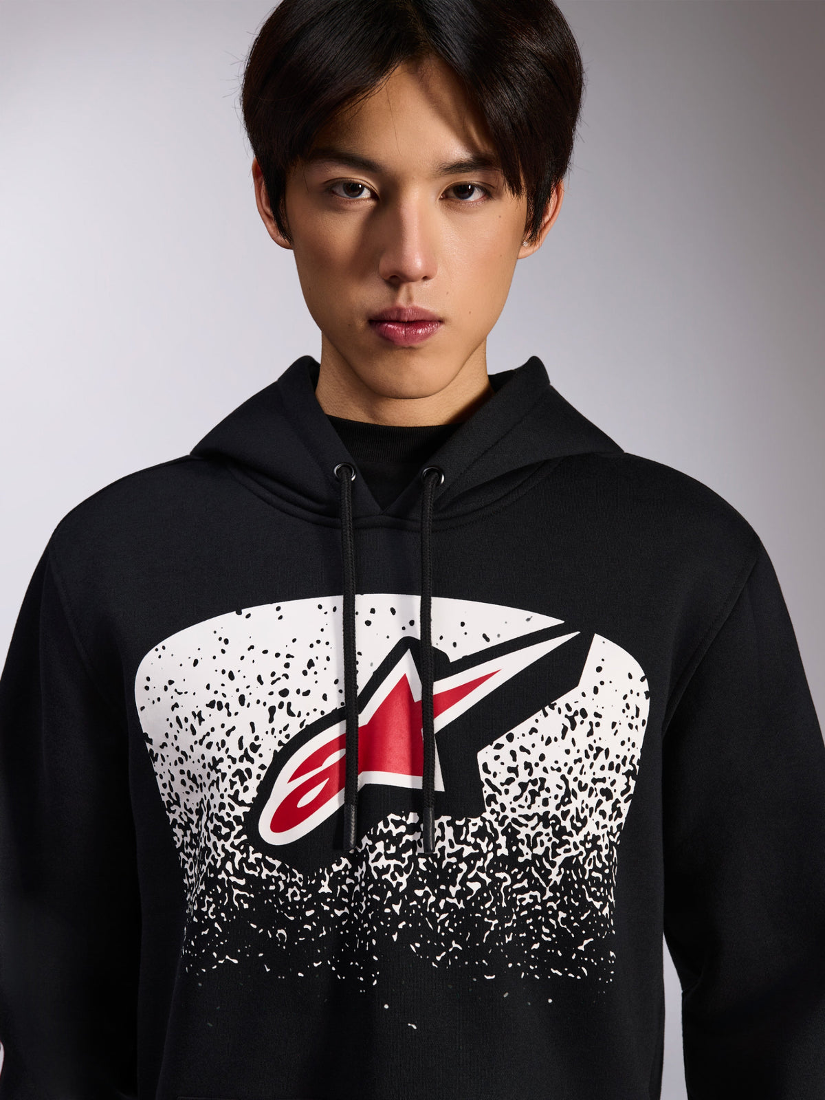 Alpinestars Obsessed Hoodie, black pullover fleece, featuring a red and white Astars logo inside a white stippled MX gradation graphic on the chest, worn by a model, casual motorsport streetwear with adjustable drawstring hood