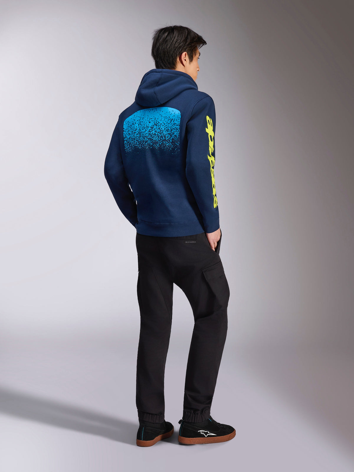 Sweat à capuche Alpinestars Obsessed, sweat-shirt décontracté bleu marine, vue de dos montrant un graphique dégradé MX bleu clair et un logo Alpinestars jaune sur la manche, porté avec un pantalon noir et des baskets