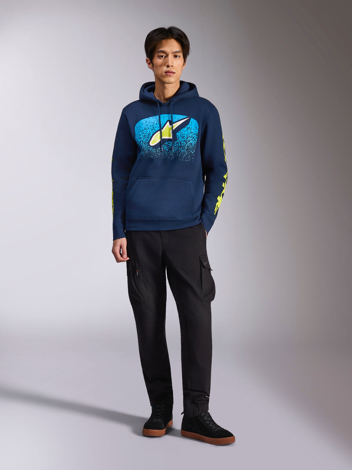 Sweat à capuche Alpinestars Obsessed, sweat bleu marine, logo Astars à l'avant avec dégradé MX bleu et étoile jaune citron, logos Alpinestars jaune citron sur les manches, porté par un mannequin avec pantalon cargo noir et baskets noires, style streetwear décontracté inspiré du sport automobile