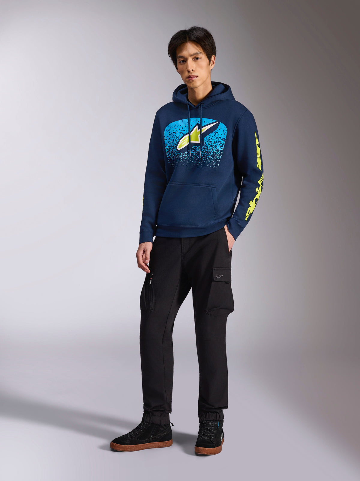 Sweat à capuche Alpinestars Obsessed en bleu marine, polaire décontractée à enfiler avec motif graphique bleu clair et jaune citron à l'avant, logo Alpinestars jaune citron sur la manche, porté par un mannequin avec un pantalon cargo noir et des baskets noires, streetwear inspiré par l'univers du sport automobile