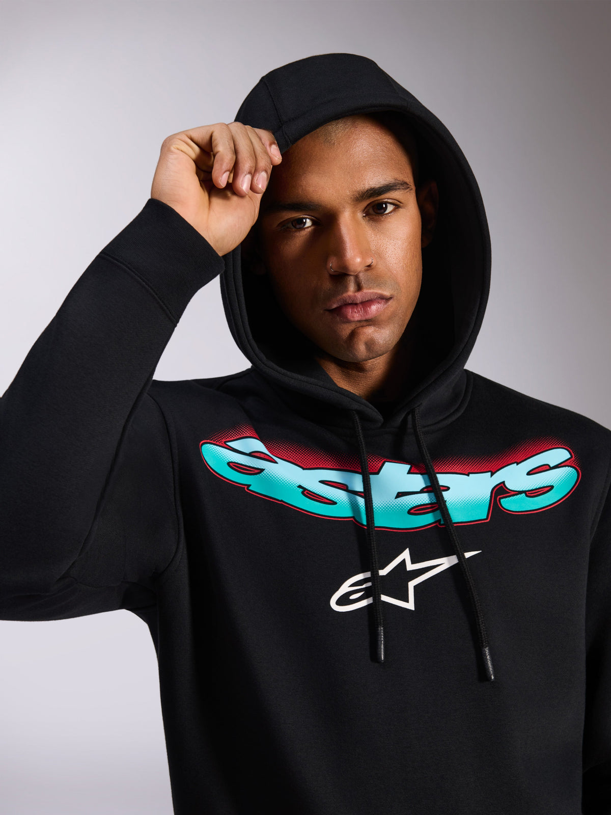 Alpinestars Ellipsoid Hoodie, black pullover fleece hoodie, featuring stylized teal and red color gradient Astars wordmark and white logo on chest, worn by model with hood up, casual motorsport-inspired streetwear