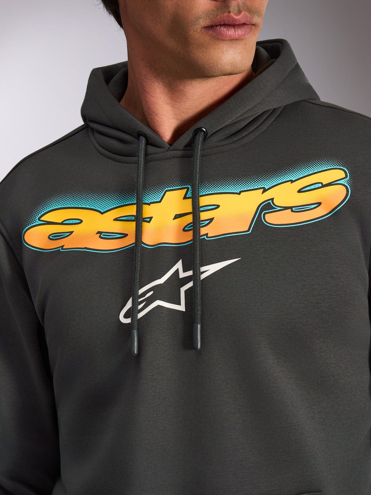 Sweat à capuche Alpinestars Ellipsoid, gris Gunmetal, gros plan sur la poitrine présentant le lettrage 'astars' incurvé avec un dégradé du jaune à l'orange et des accents bleu sarcelle, logo étoile Alpinestars blanc, polaire streetwear décontractée inspirée du sport automobile