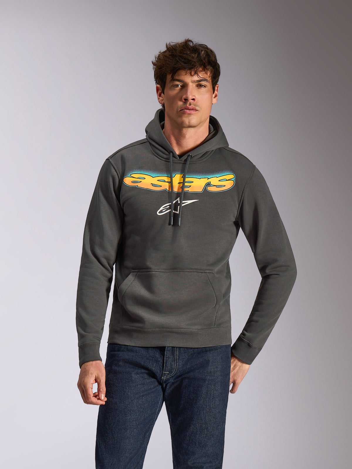 Sweat à capuche Alpinestars Ellipsoid, vêtement de sport décontracté, gris Gunmetal avec lettrage Astars en dégradé jaune et bleu et logo blanc, modèle à enfiler avec capuche à cordon de serrage, porté avec un jean bleu foncé