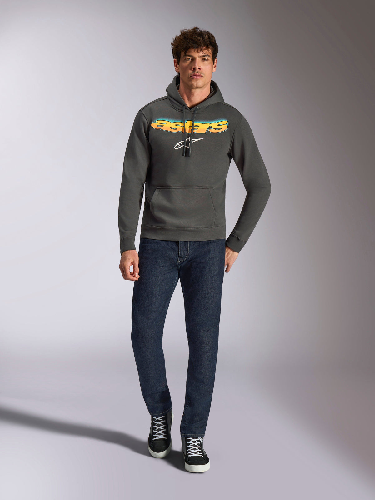 Sweat à capuche Alpinestars Ellipsoid, sportswear décontracté, gris anthracite avec logo Astars en dégradé orange et jaune et symbole blanc, capuche avec cordon de serrage, porté par un mannequin homme avec un jean bleu foncé et des baskets noires, streetwear moderne inspiré du sport automobile