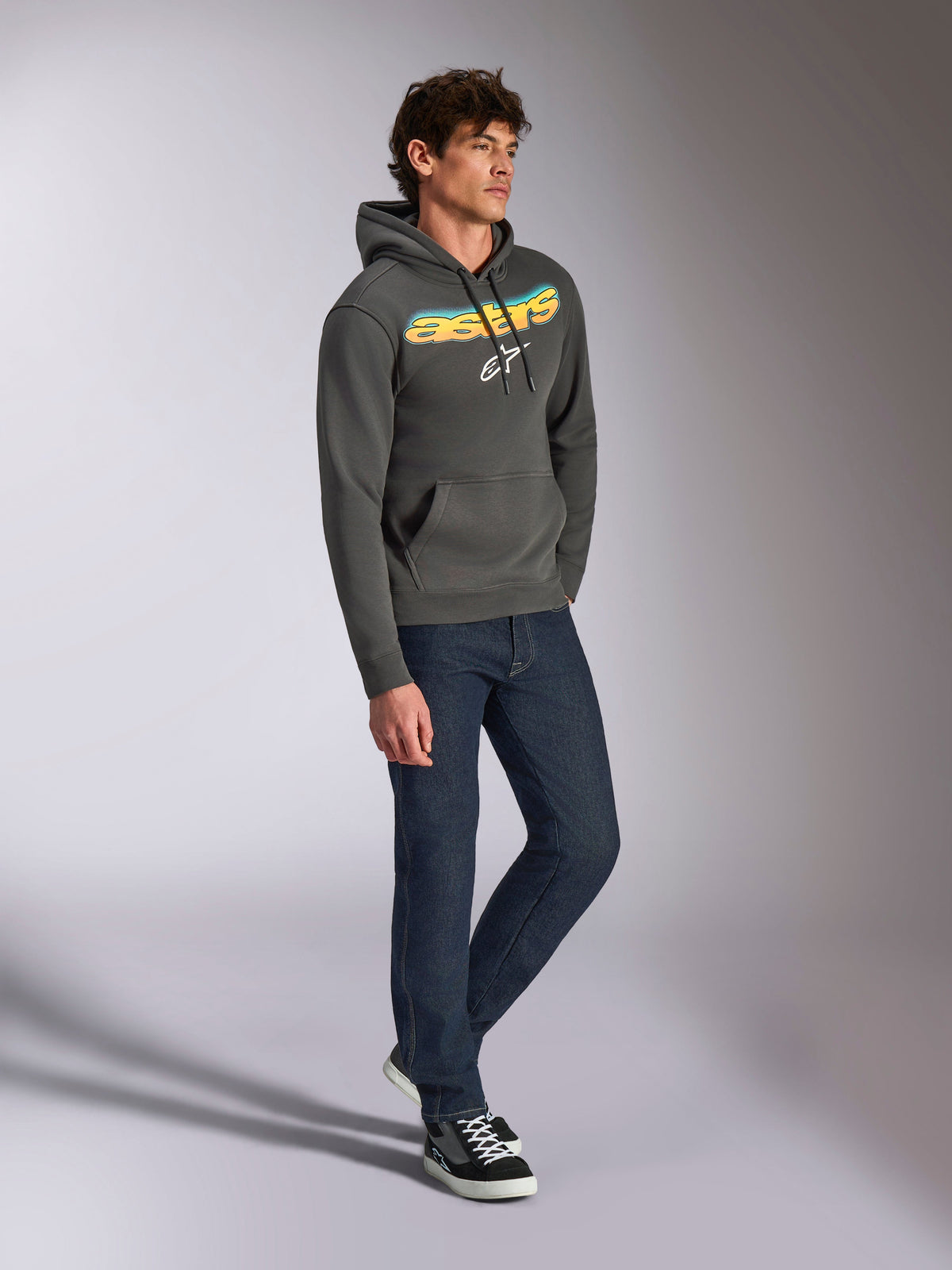 Sweat à capuche Alpinestars Ellipsoid, polaire à enfiler gris anthracite, lettrage Astars incurvé à l'avant avec dégradé bleu et jaune et logo blanc, porté avec un jean foncé et des baskets noires, style streetwear décontracté.