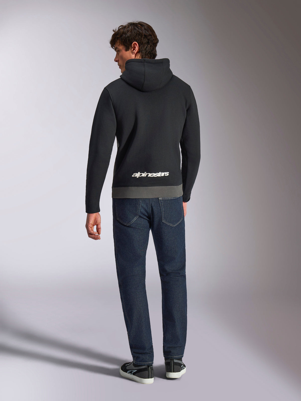Sweat à capuche Parallel Alpinestars, vue de dos d'un homme portant un sweat en polaire à enfiler noir et anthracite, logo Alpinestars blanc dans le bas du dos, associé à un jean bleu foncé, style streetwear décontracté inspiré des sports mécaniques