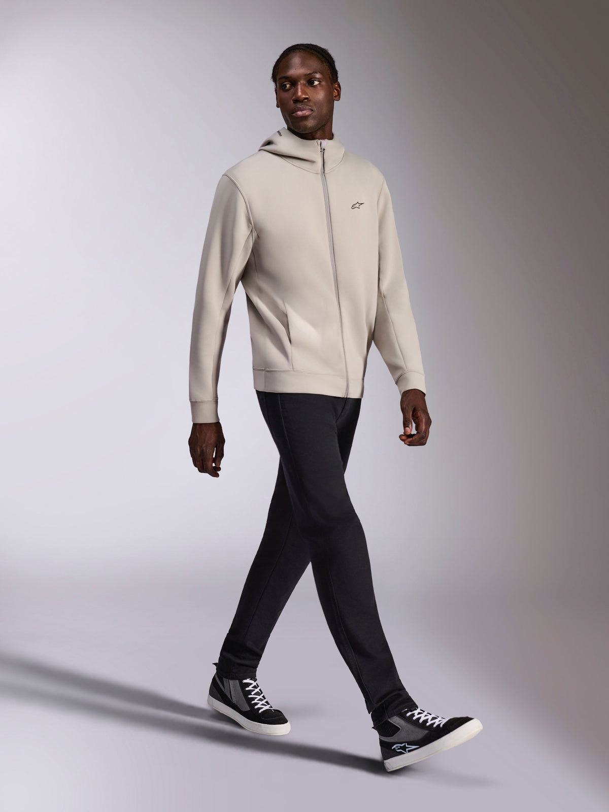 Sweat à capuche Alpinestars Cypher, polaire zippée gris ciment, capuche à col haut, petit logo Astars sur la poitrine, porté par un mannequin en train de marcher, associé à un pantalon noir et des baskets noires et grises, style sportswear décontracté d'inspiration motorsport.