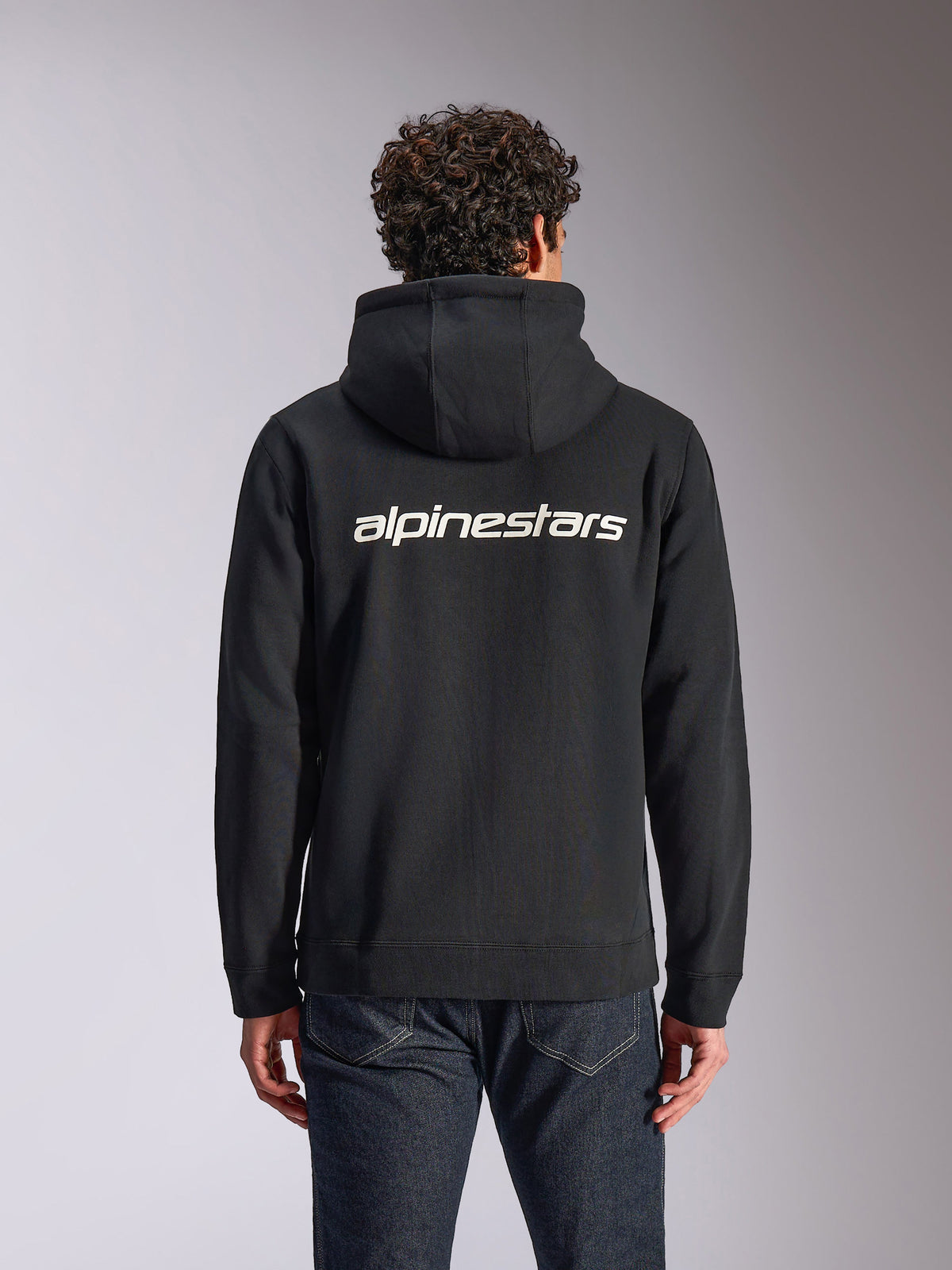 Sweat à capuche Alpinestars Transverse, style streetwear décontracté, noir avec logo Alpinestars blanc dans le dos, polaire à capuche avec fermeture éclair frontale, manches longues, porté par un mannequin avec un jean en denim foncé, design moderne inspiré du sport automobile