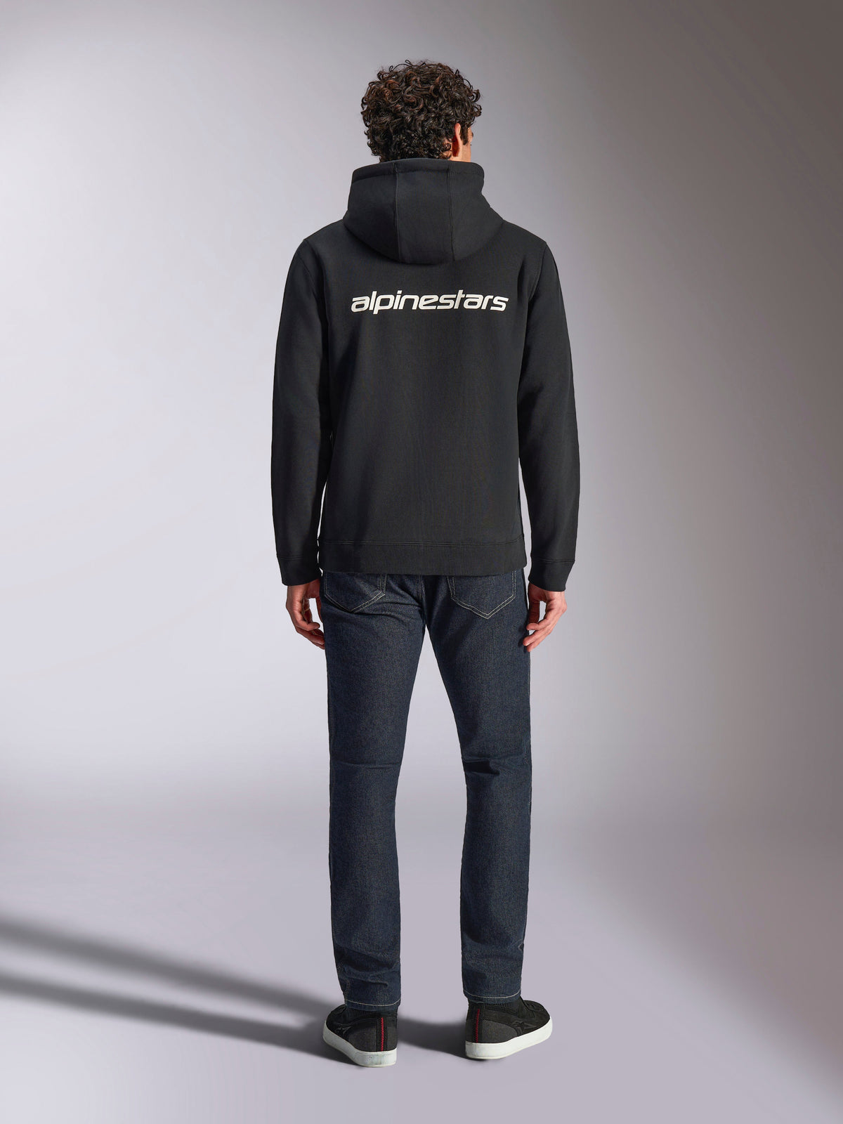 Sweat à capuche Alpinestars Transverse, streetwear motorsport décontracté, noir avec logo Alpinestars blanc au dos, manches longues, ourlet et poignets côtelés, vu de dos avec un jean en denim foncé