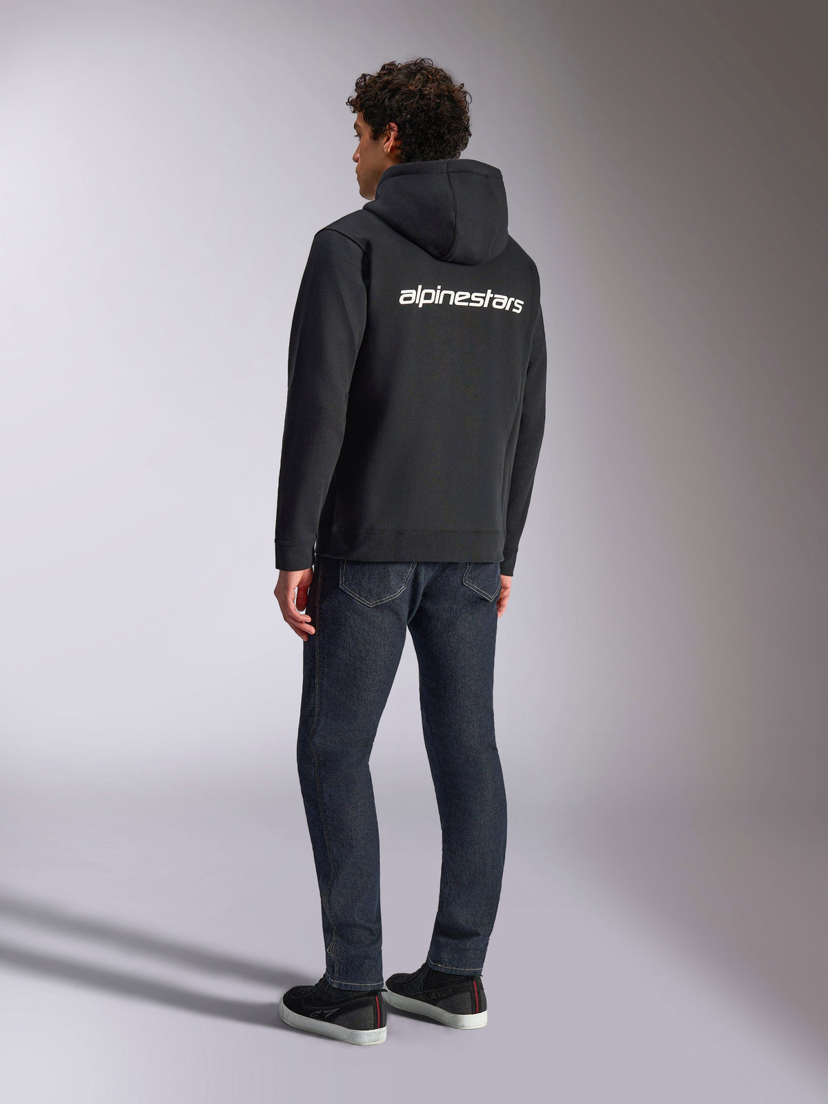 Sweat à capuche Alpinestars Transverse, polaire noire avec fermeture éclair, vue de dos montrant un logo Alpinestars blanc sur les épaules, porté avec un jean bleu foncé et des baskets noires, streetwear décontracté inspiré des sports mécaniques
