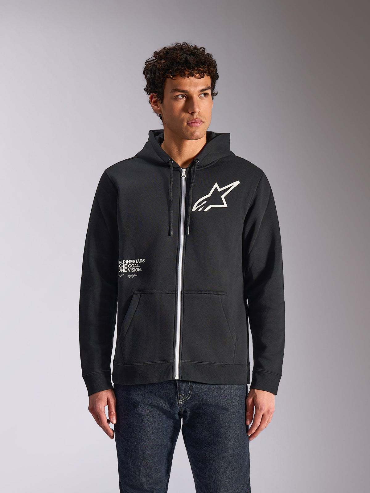 Sweat à capuche Alpinestars Transverse, polaire noire à fermeture éclair frontale, avec logo Astars blanc et motif One Goal sur le devant, porté par un mannequin avec un jean foncé, look streetwear décontracté inspiré du sport automobile
