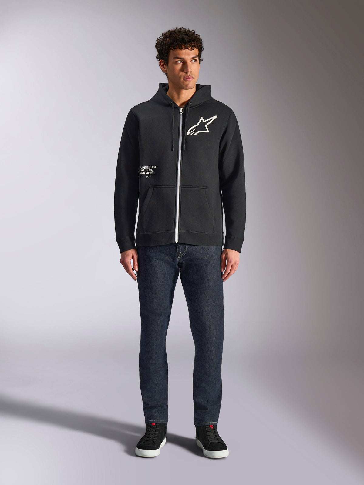 Sweat à capuche Alpinestars Transverse, polaire décontractée à fermeture éclair frontale, noir, logo Astars blanc sur la poitrine et imprimé 'One Goal' sur le devant, capuche à cordon de serrage, associé à un jean foncé et des baskets noires, style streetwear sport automobile