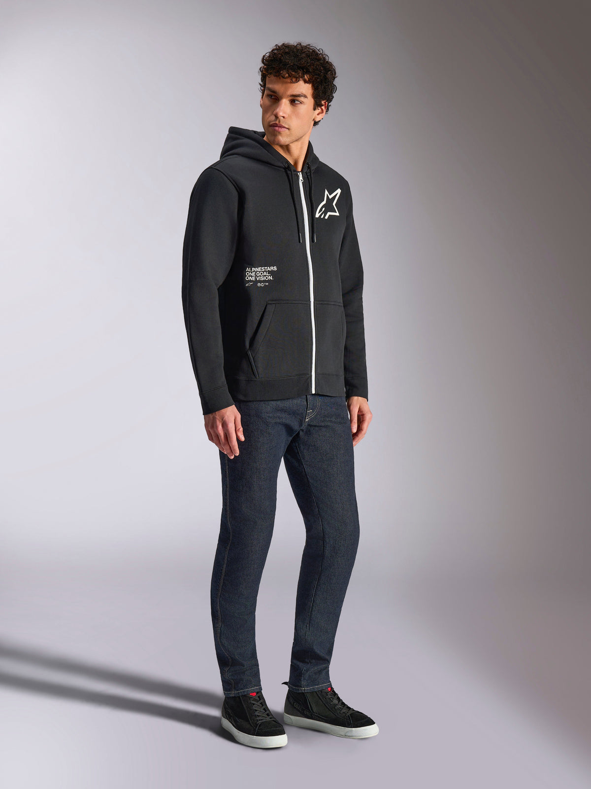 Sweat à capuche Alpinestars Transverse, hoodie zippé noir avec fermeture éclair blanche contrastante, logo étoile blanc sur la poitrine et marquage « One Goal » sur la poche, porté par un mannequin avec un jean foncé et des baskets noires, streetwear décontracté inspiré du sport automobile.