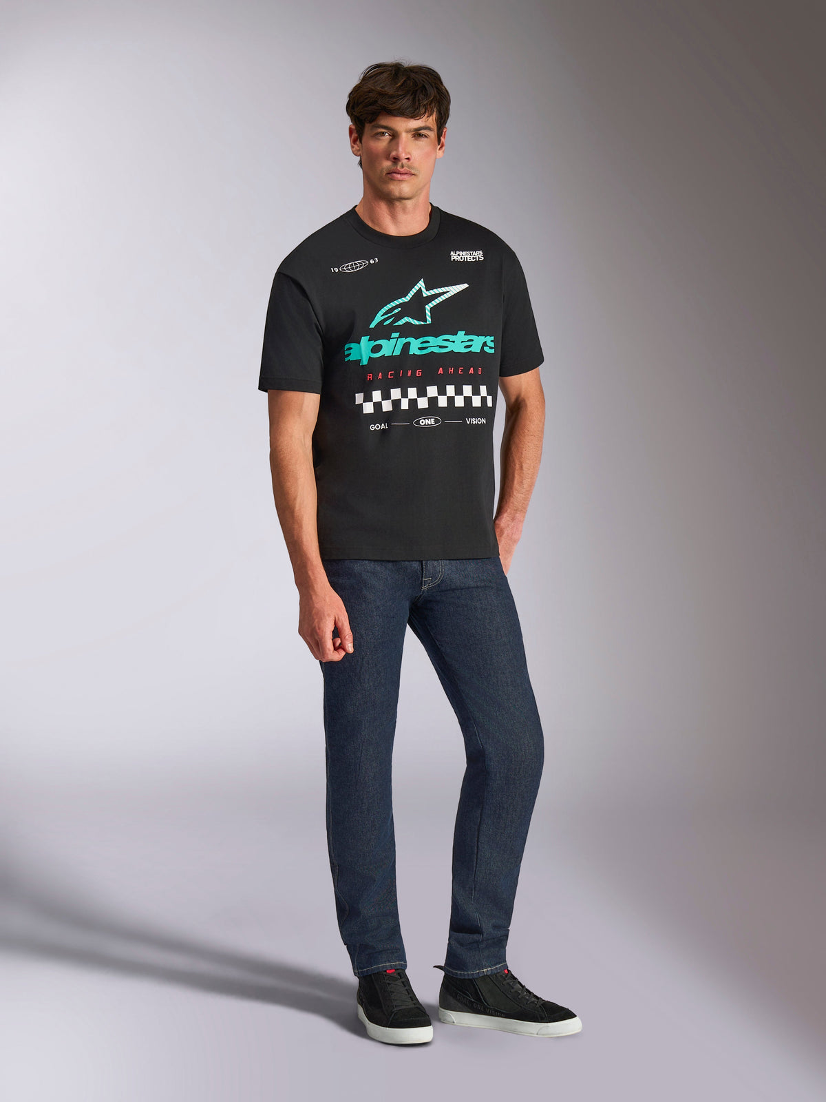 T-shirt Aplomb CSF Oversized Alpinestars, t-shirt à manches courtes, noir, grand logo Alpinestars blanc et bleu sarcelle avec dégradé, texte « Racing Ahead » et motif à damier, porté avec un jean bleu foncé et des baskets noires, style streetwear motorsport décontracté