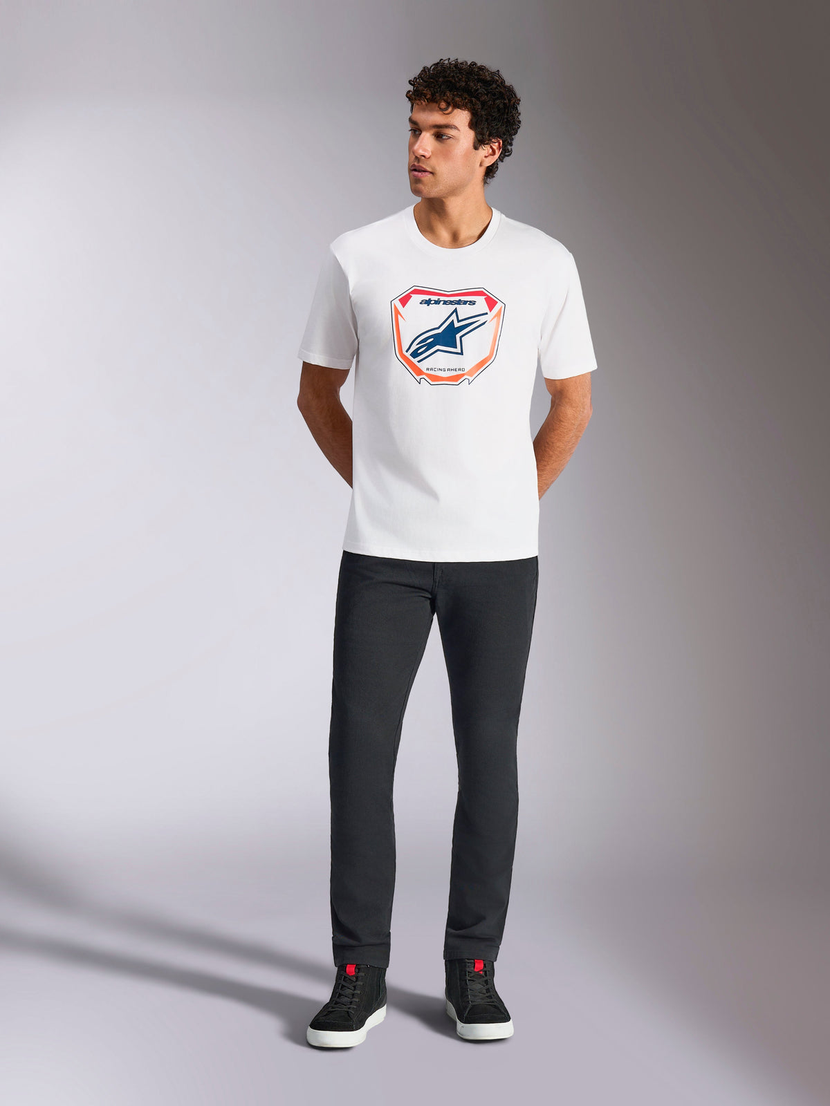 T-shirt Alpinestars Plate CSF, t-shirt à manches courtes, blanc, arborant un graphique de plaque Astars MX à l'avant avec des accents orange et bleu, porté par un mannequin avec un pantalon noir et des baskets, style streetwear décontracté