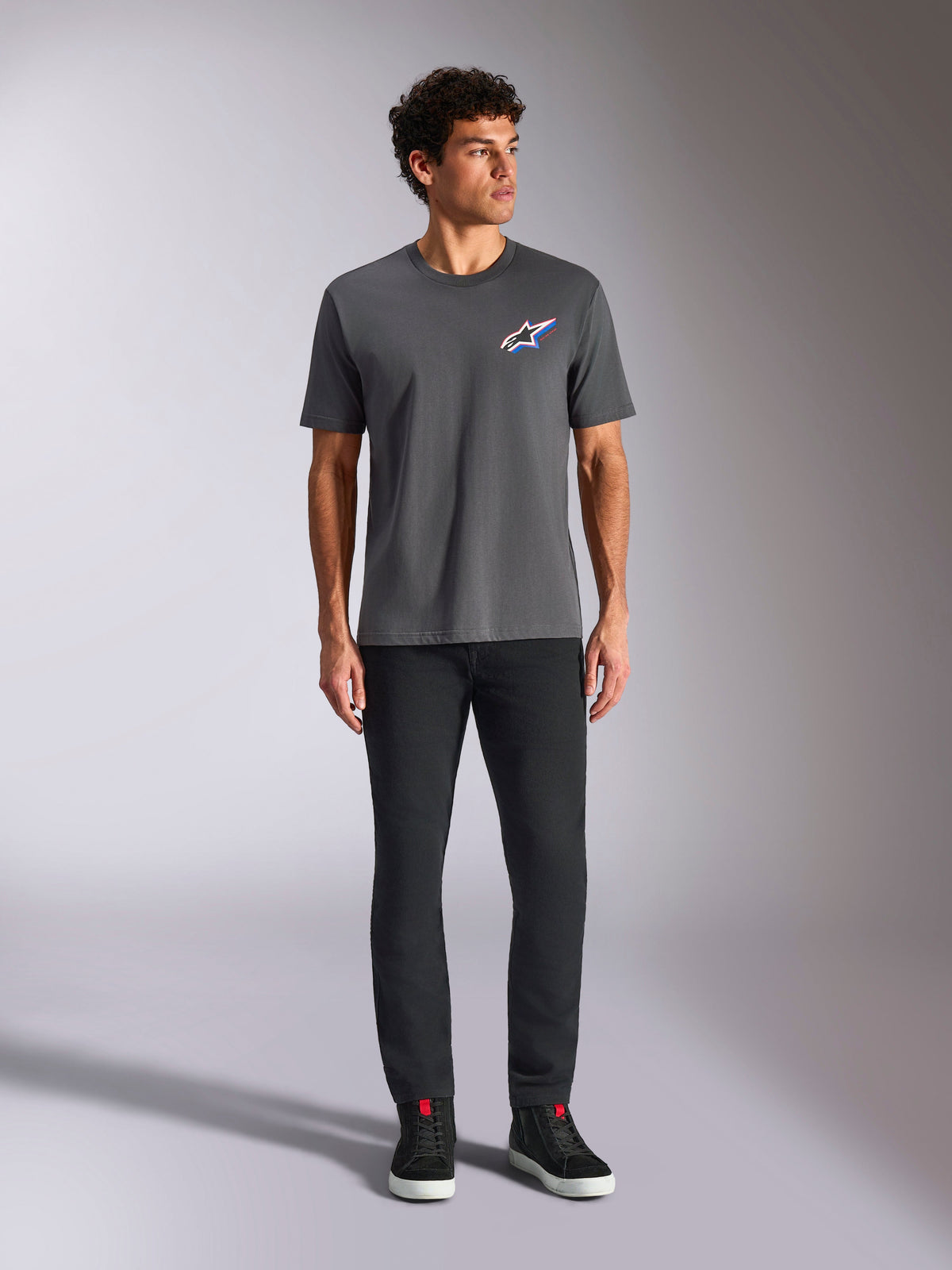 T-shirt Alpinestars Angler CSF, t-shirt à manches courtes, anthracite, avec un logo Astars multicolore éclatant sur la poitrine, porté par un mannequin avec un pantalon noir et des baskets, style streetwear décontracté motorsport
