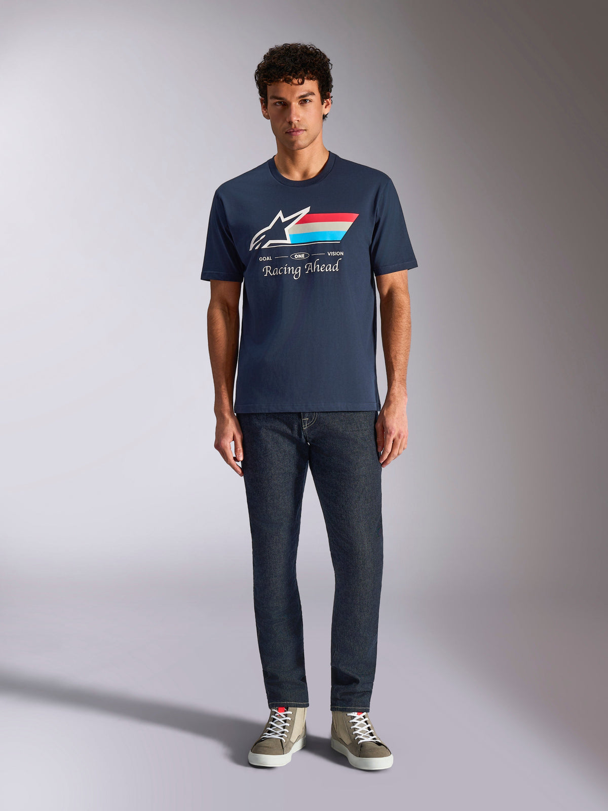 Alpinestars Trifecta CSF Tee, t-shirt à manches courtes, bleu marine, logo Astars blanc avec rayures rouges, blanches et bleu clair et texte « Racing Ahead », porté avec un jean foncé et des baskets olive, style streetwear motorsport décontracté