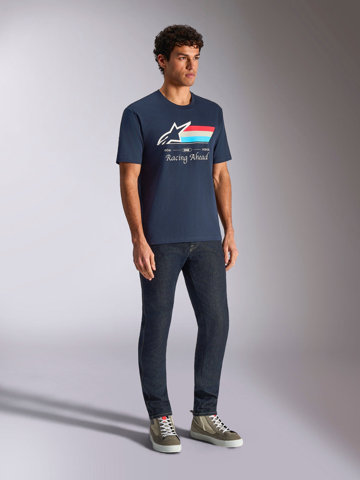 T-shirt Alpinestars Trifecta CSF, manches courtes, bleu marine, avec logo Astars, rayures tricolores et texte « Racing Ahead », porté par un mannequin avec un jean foncé et des baskets, style streetwear décontracté inspiré du sport automobile