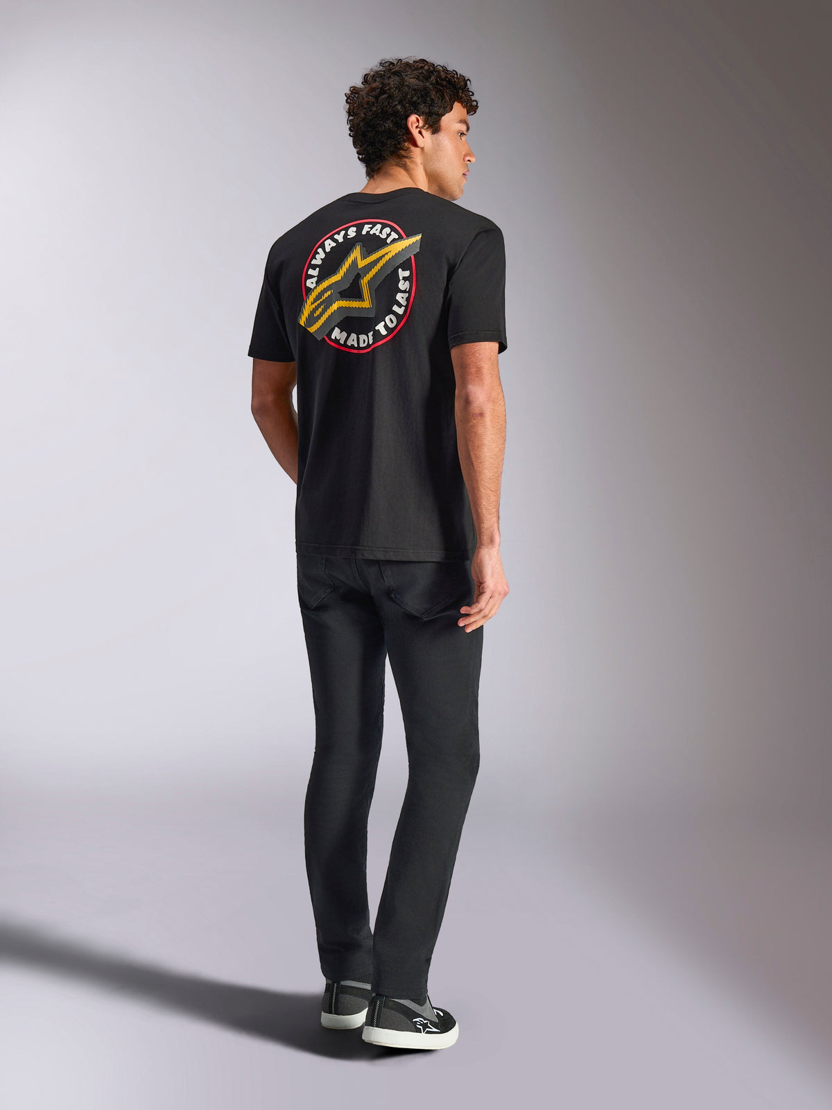 T-shirt à manches courtes Alpinestars Aorbit CSF, noir, vue de dos présentant un grand logo Astars jaune avec le motif circulaire 'Always Fast Made to Last', porté avec un jean noir et des baskets décontractées, style streetwear inspiré du sport automobile