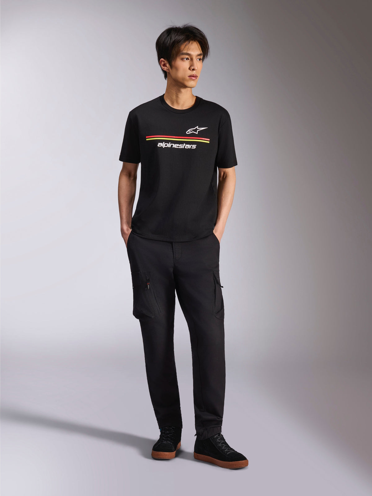 Modèle portant le t-shirt Alpinestars Lanes CSF, t-shirt à manches courtes noir avec des rayures rouges et jaunes et les logos Astars et Alpinestars, associé à un pantalon cargo noir et des baskets, style streetwear de sport automobile décontracté