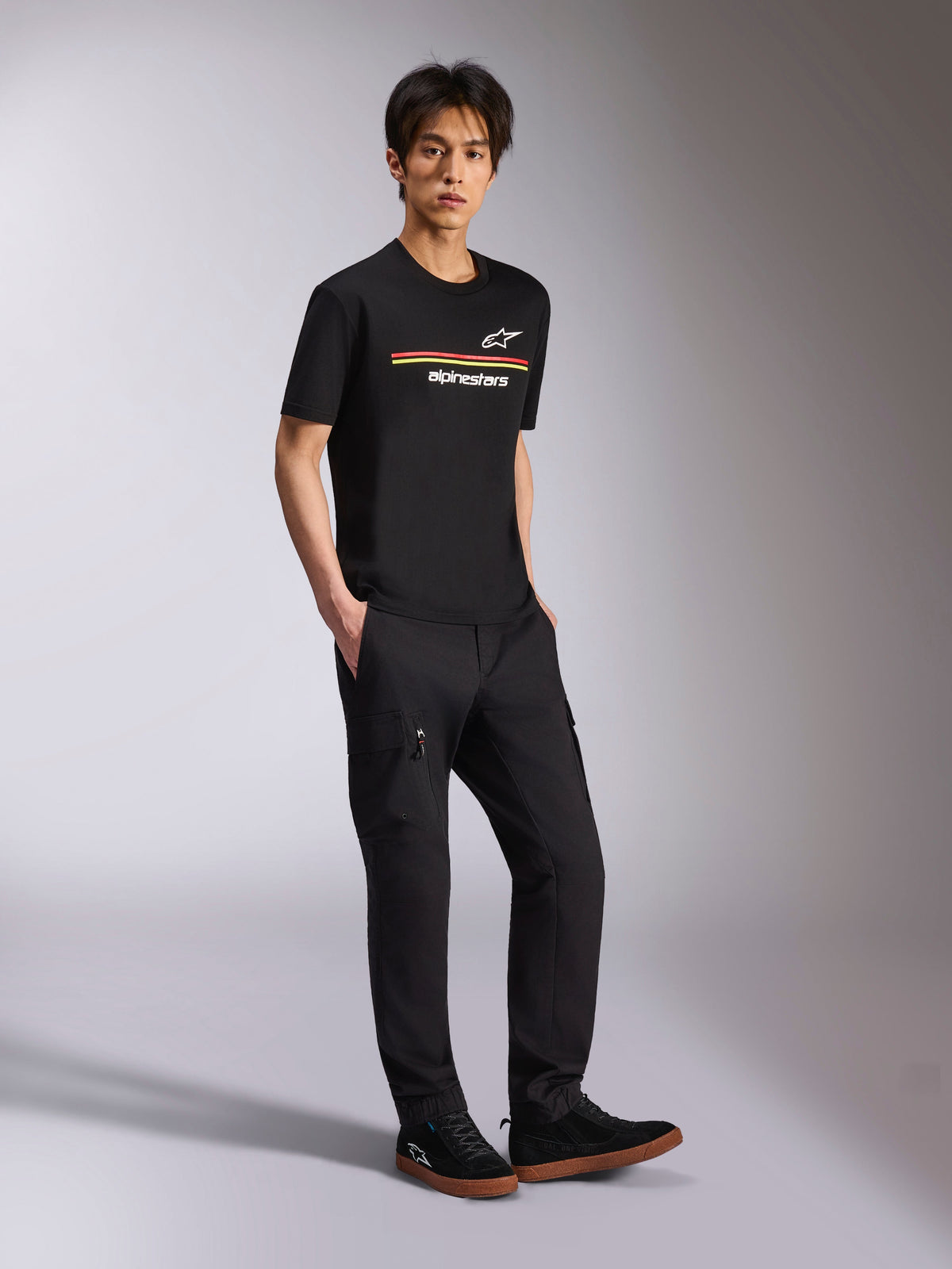 T-shirt à manches courtes Alpinestars Lanes CSF, noir, logos Astars et Alpinestars avec rayures multicolores sur la poitrine, porté par un mannequin avec un pantalon cargo noir, streetwear décontracté