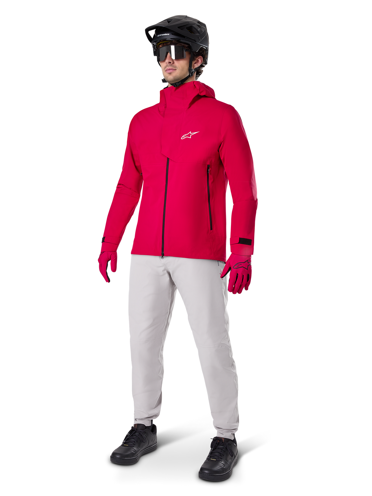 Cycliste portant la veste imperméable Alpinestars A-Dura Elite en Rouge Fluo, associée à un pantalon technique gris clair, des gants rouges, un casque de VTT noir avec lunettes et des chaussures noires, tenue haute performance complète pour le VTT